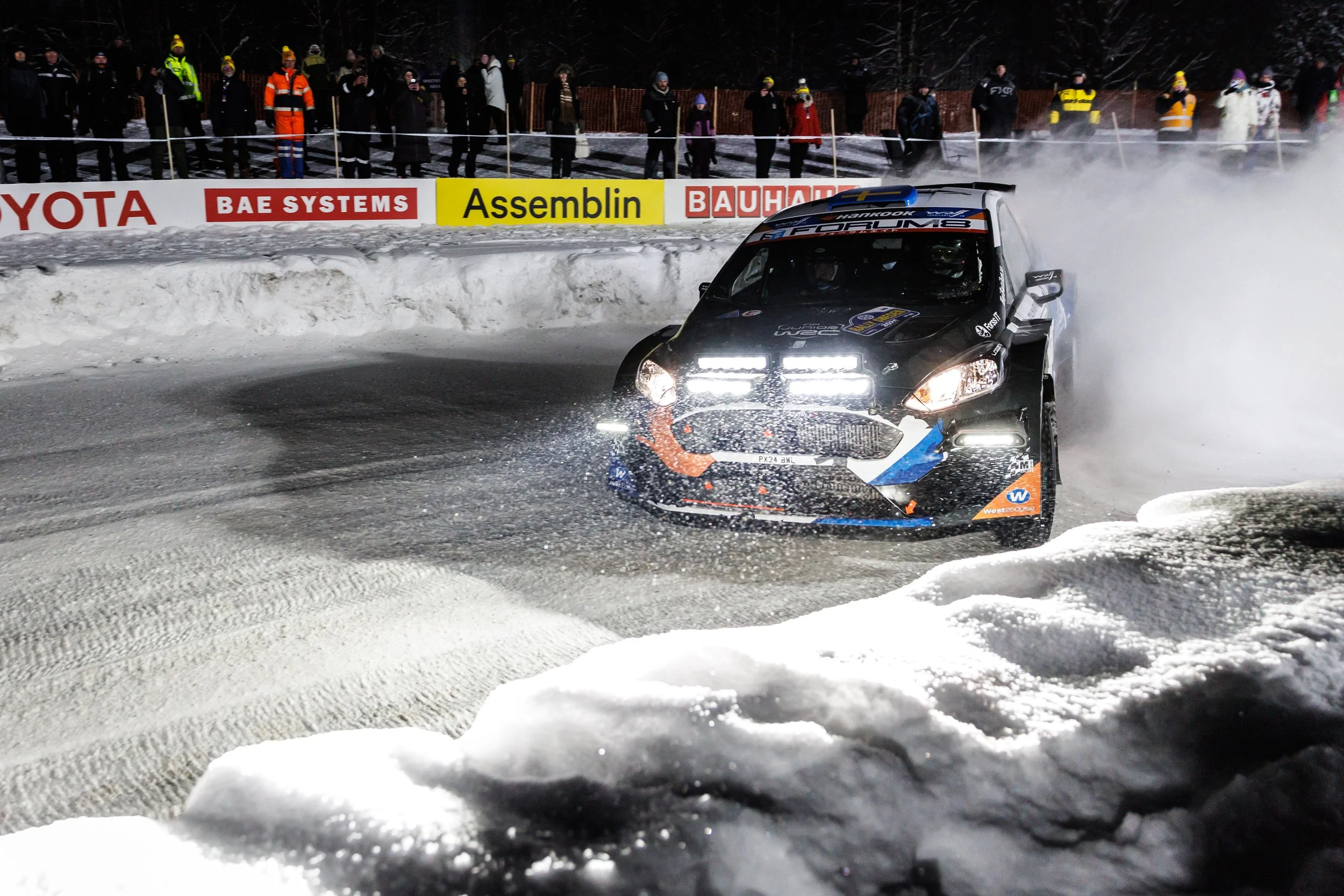 A black and blue rally car racing on a snowy track at night, with snow being kicked up behind it. Spectators in winter clothing stand behind safety barriers along the track, and advertisements are visible on the barriers.