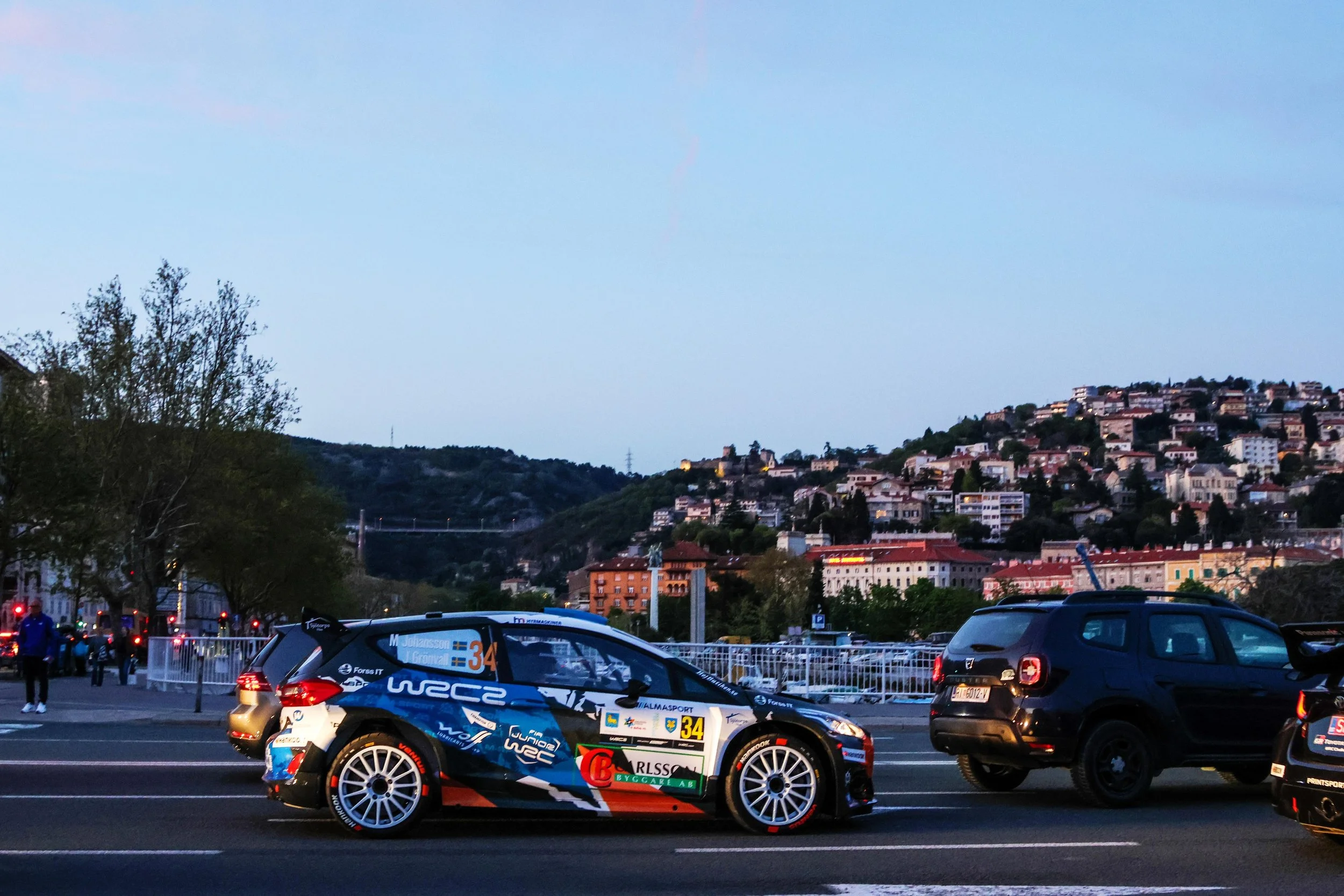 A rally car with racing decals and sponsor logos parked in a parking lot at dusk, with a hillside town and a blue sky in the background.