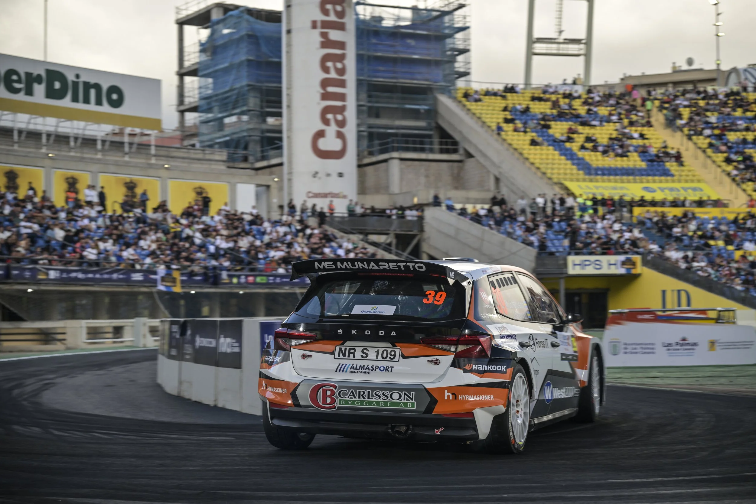 A rally car racing on a track with a crowd of spectators in the background, surrounded by barriers and advertisements.