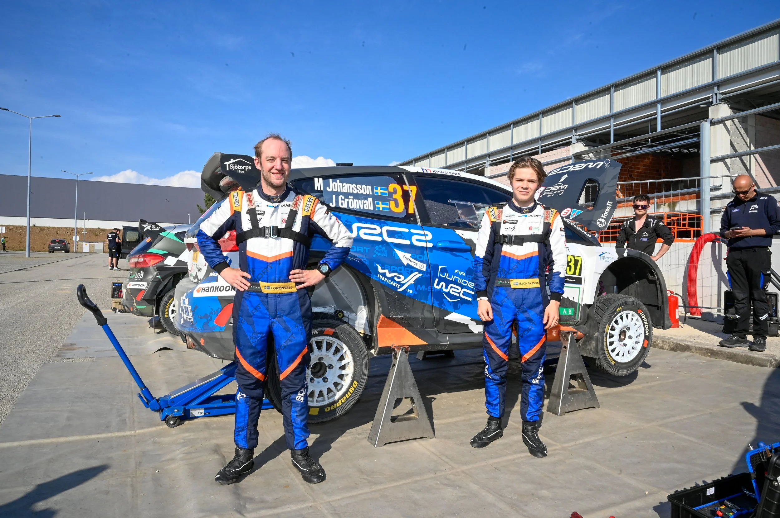 Two rally drivers in racing suits standing in front of a rally car marked with the Swedish flag and the names M. Johansson and J. Grönvall. The scene is outdoors on a sunny day, with equipment and team members nearby.