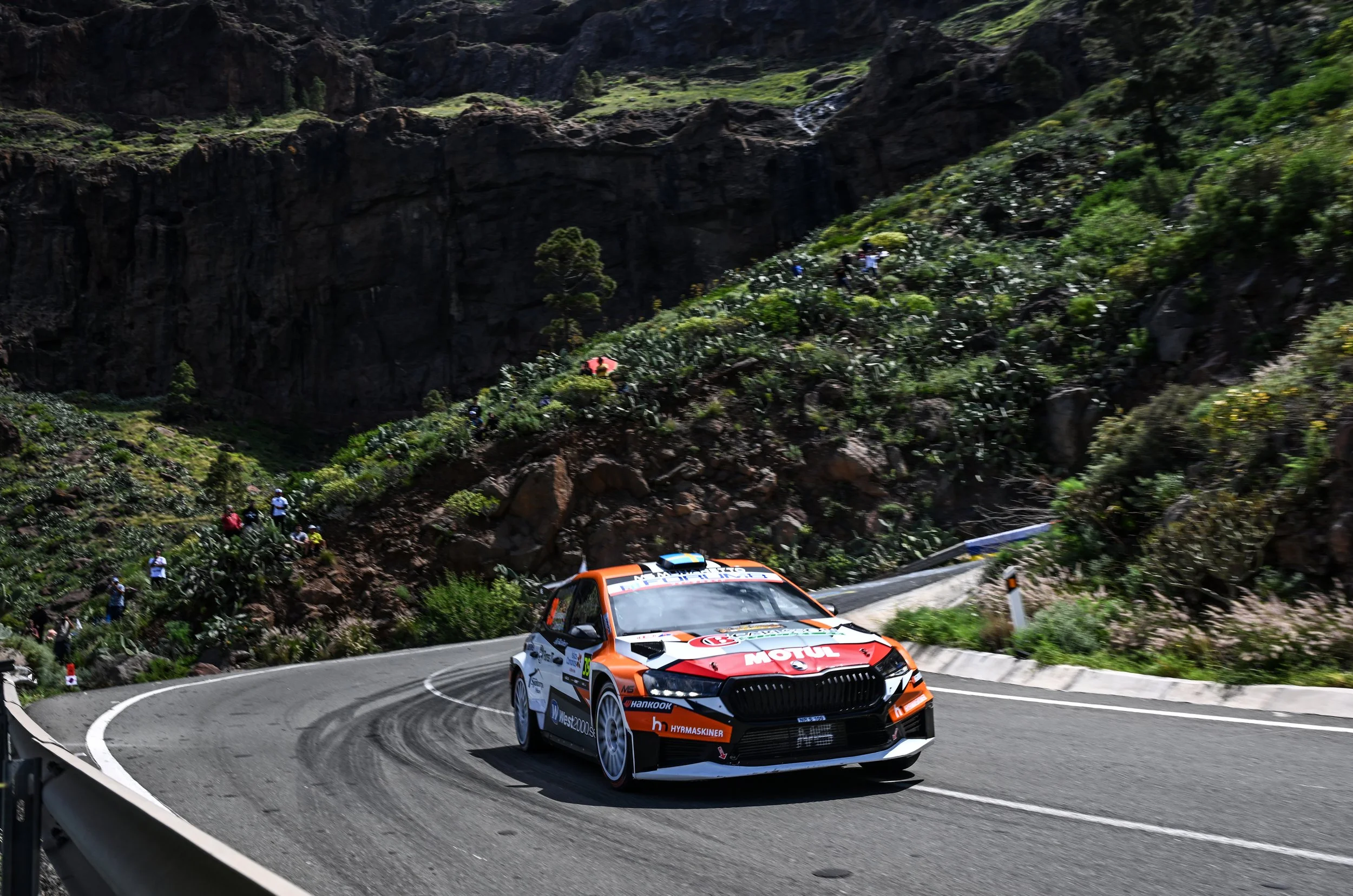 A rally car racing on a winding mountain road surrounded by rocky cliffs and green vegetation with spectators watching from the roadside.