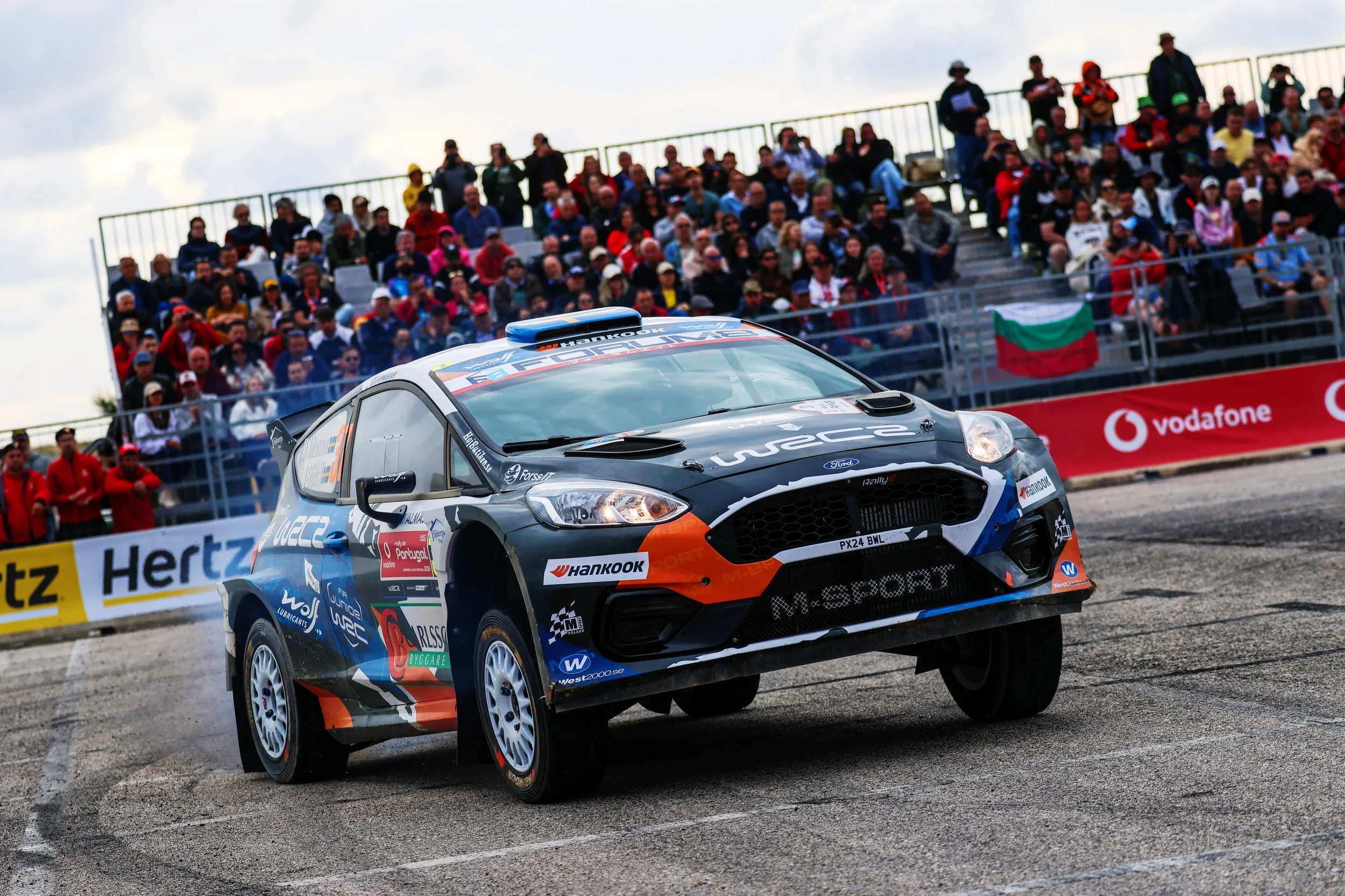 A rally car is jumping in the air on a dirt rally stage with a crowd of spectators watching from the stands in the background.