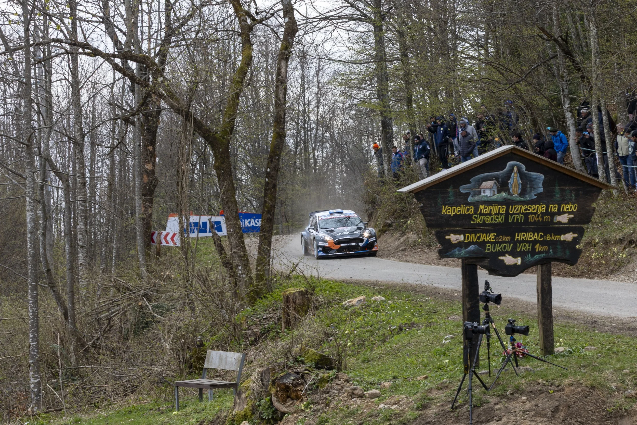 Race car on a dirt road in a forest, with a crowd of spectators above on a hillside. Signs indicate directions and distances in the forest area.