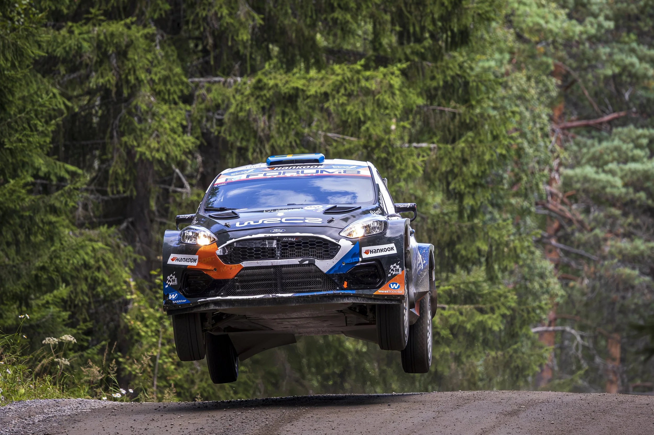A rally car flying through the air over a dirt road surrounded by green trees.