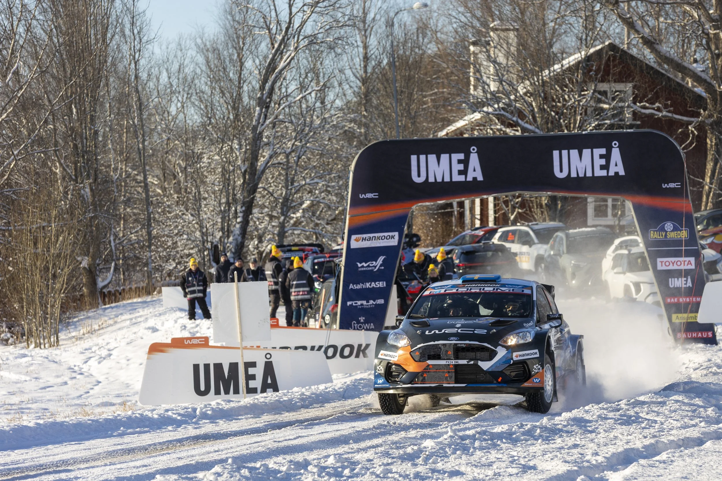 A rally car racing on a snowy track during the UMEA stage of a rally event, with spectators and crew members in winter clothing. There are banners and signage with rally branding and sponsors, and snow-covered trees and houses in the background.