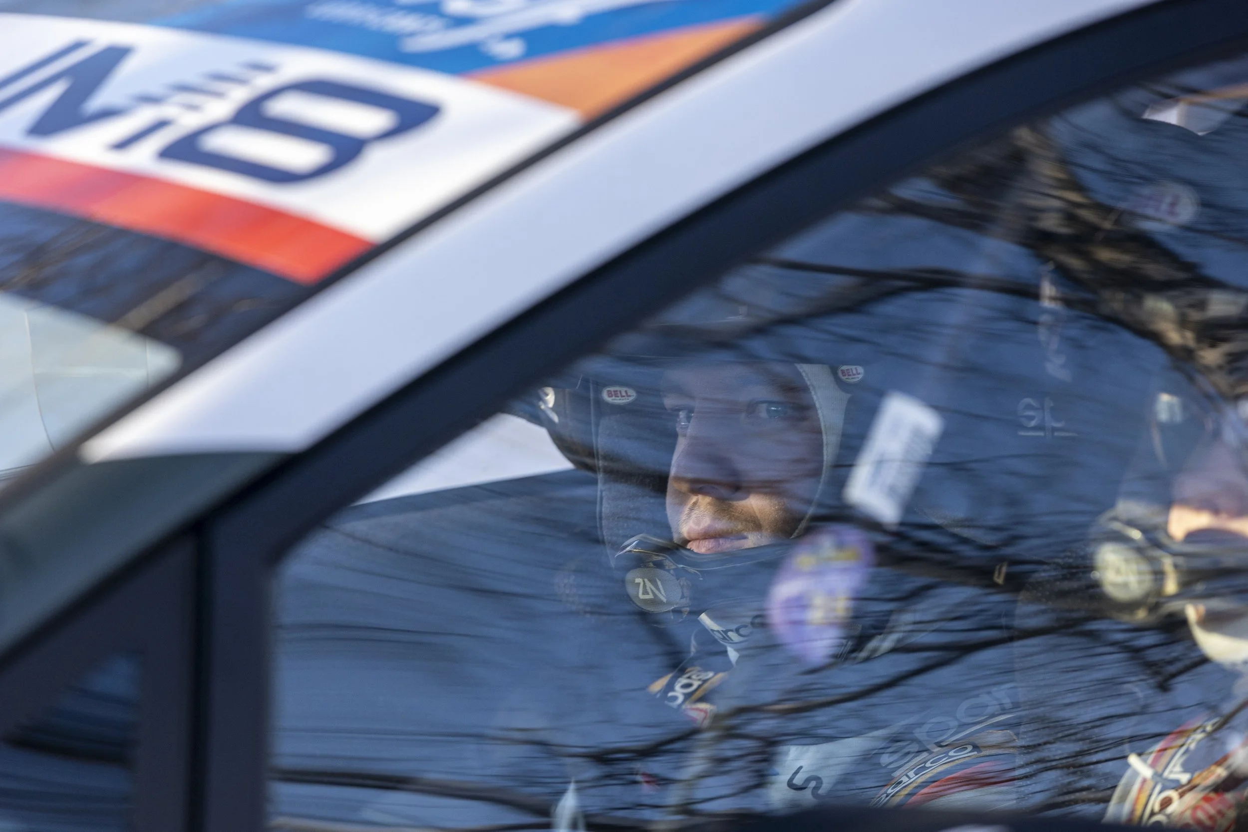 Close-up of a race car driver inside a car, wearing a helmet and racing suit, reflected in the car window, with a blurred background.