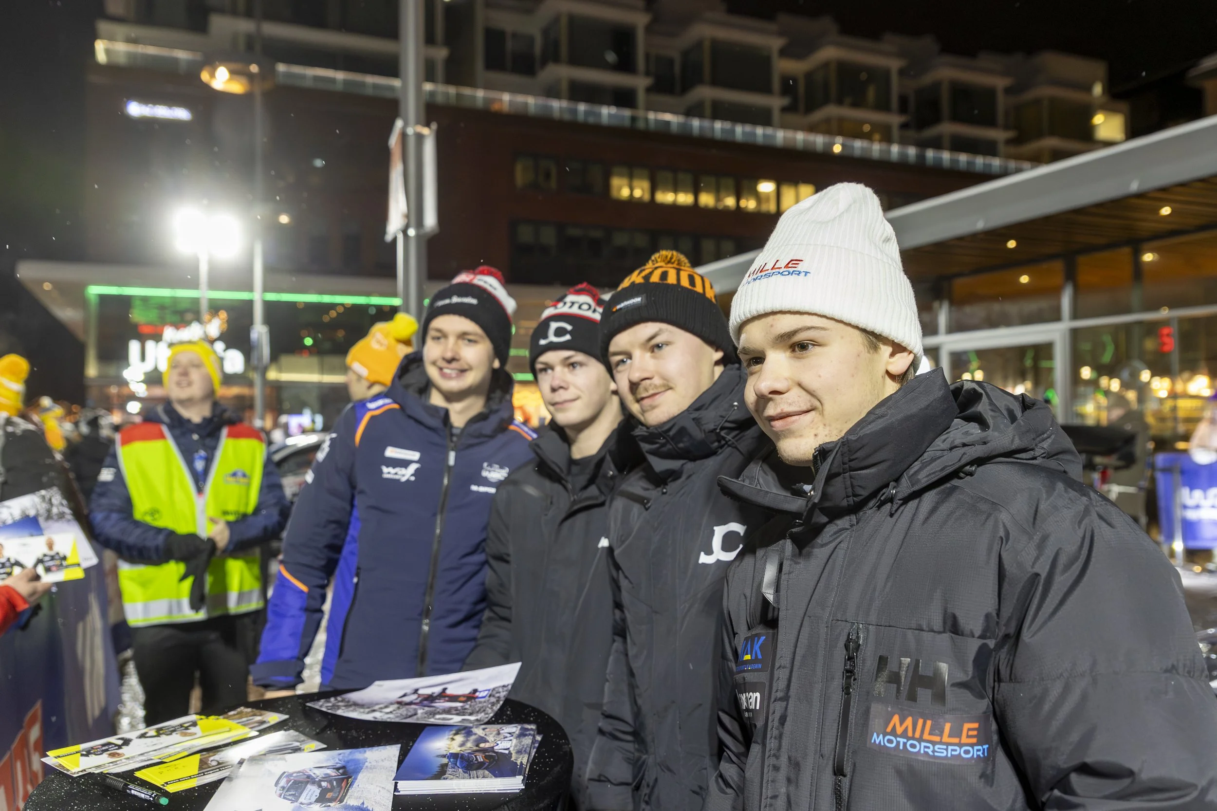 A group of young men in racing jackets and hats standing outdoors at night, likely at a motorsport event, with a building and bright lights in the background.
