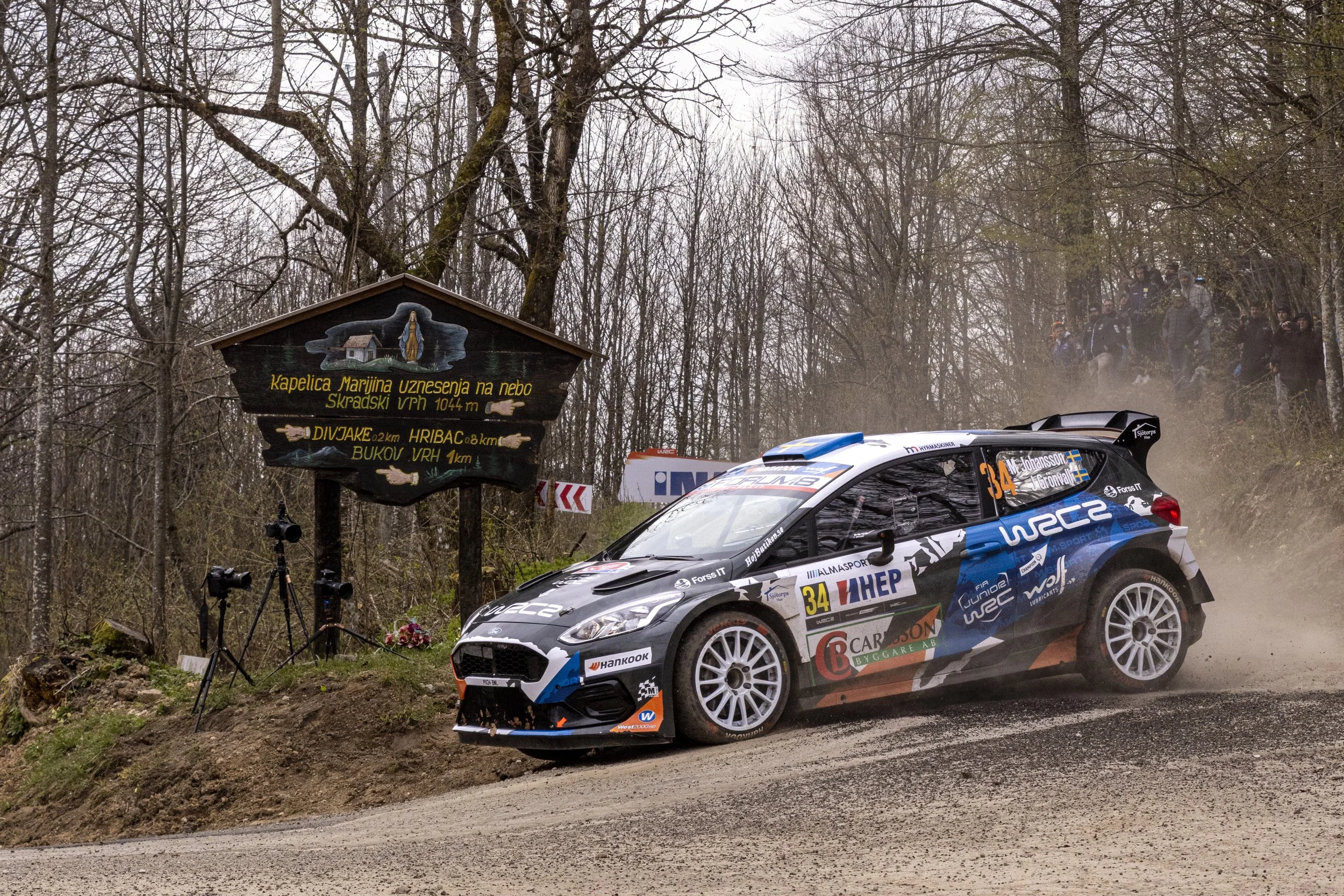 A rally car speeds along a dirt road surrounded by trees, with a rally sign and spectators in the background.