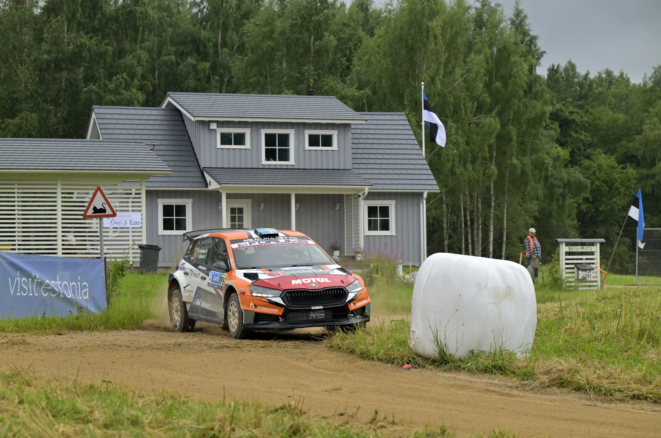 A rally car speeding on a dirt track mid-turn with dust trailing behind, with a house and trees in the background, and a person standing near a flag on the right side.