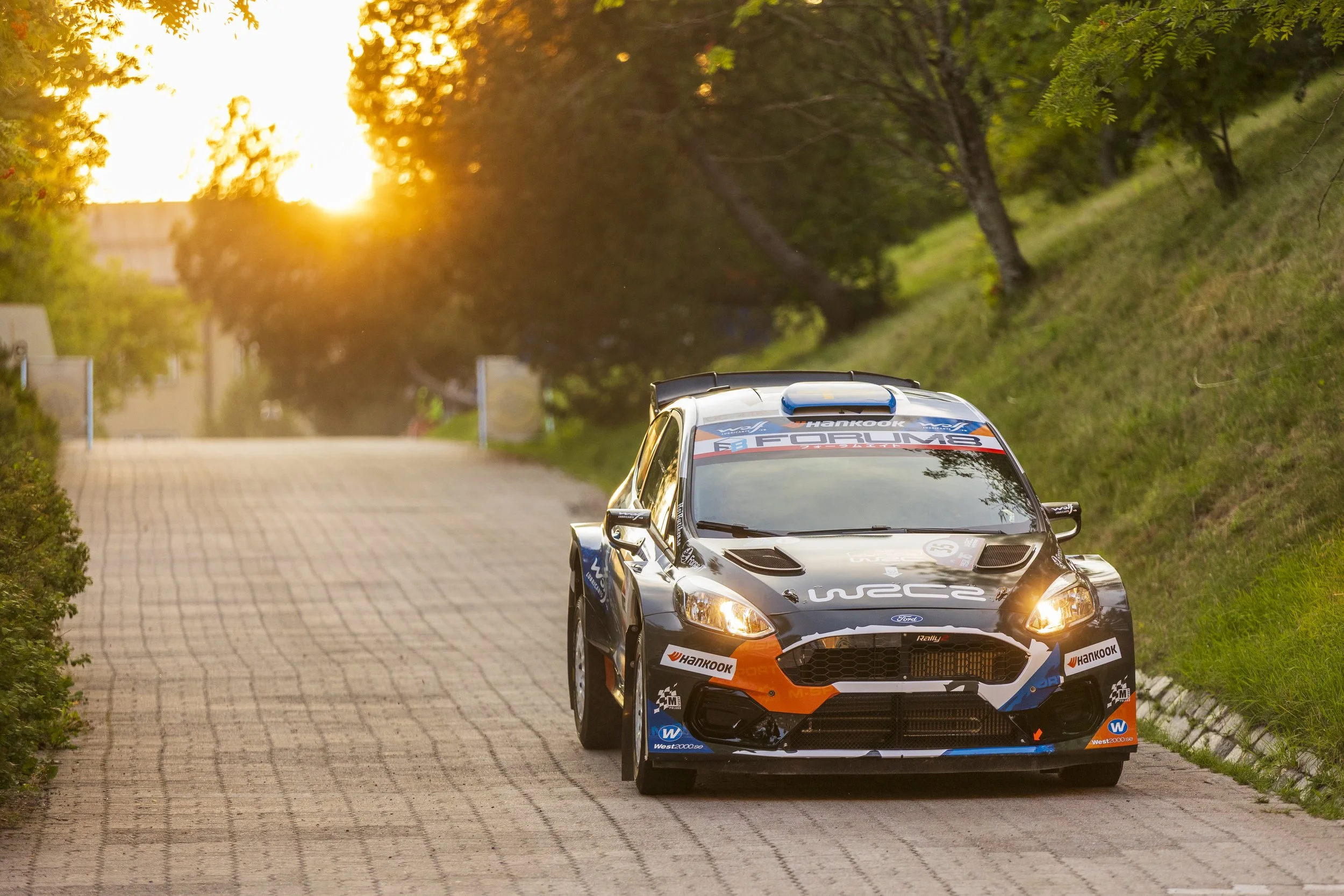 A black and orange rally car with sponsor logos, including Hankook tires, driving on a paved road lined with green grass and trees, during sunset.
