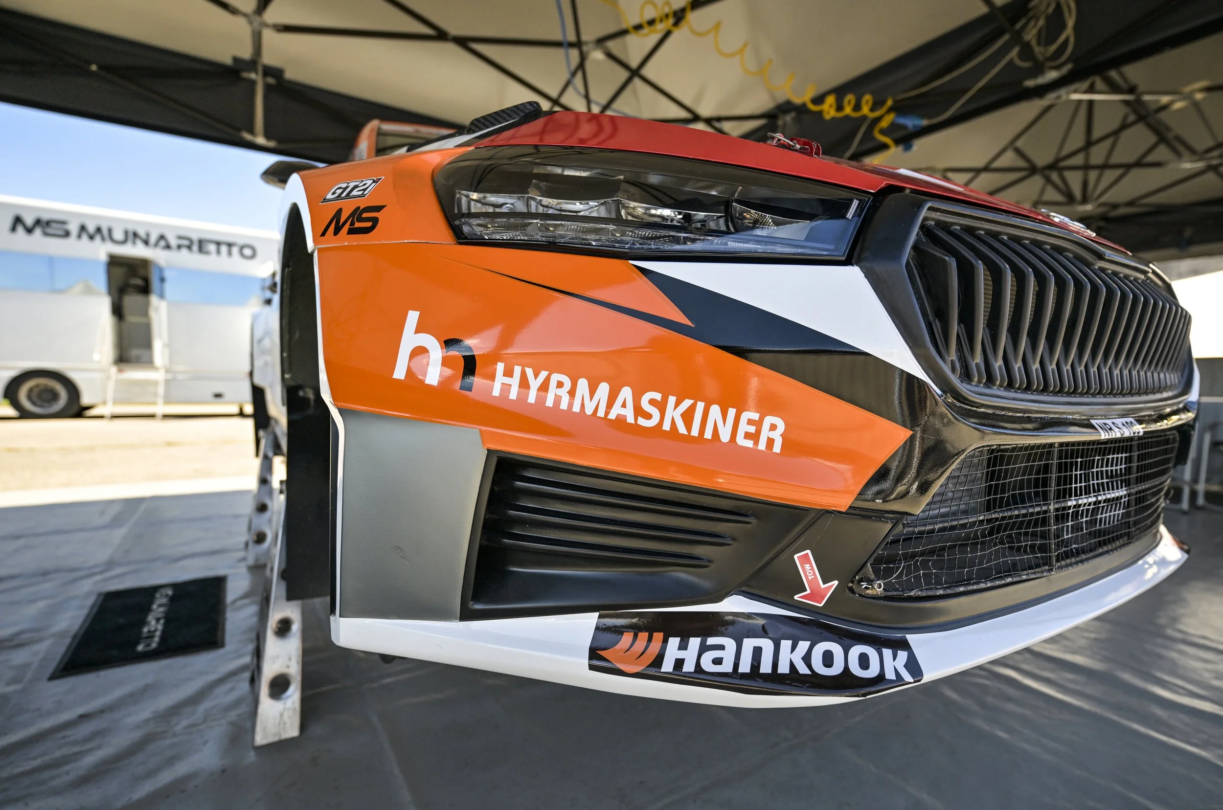 Close-up of a race car's front, showing sponsor logos such as Hankook, Hyundai, and MS. The car is in a garage with a tent above, with other vehicles and equipment visible in the background.