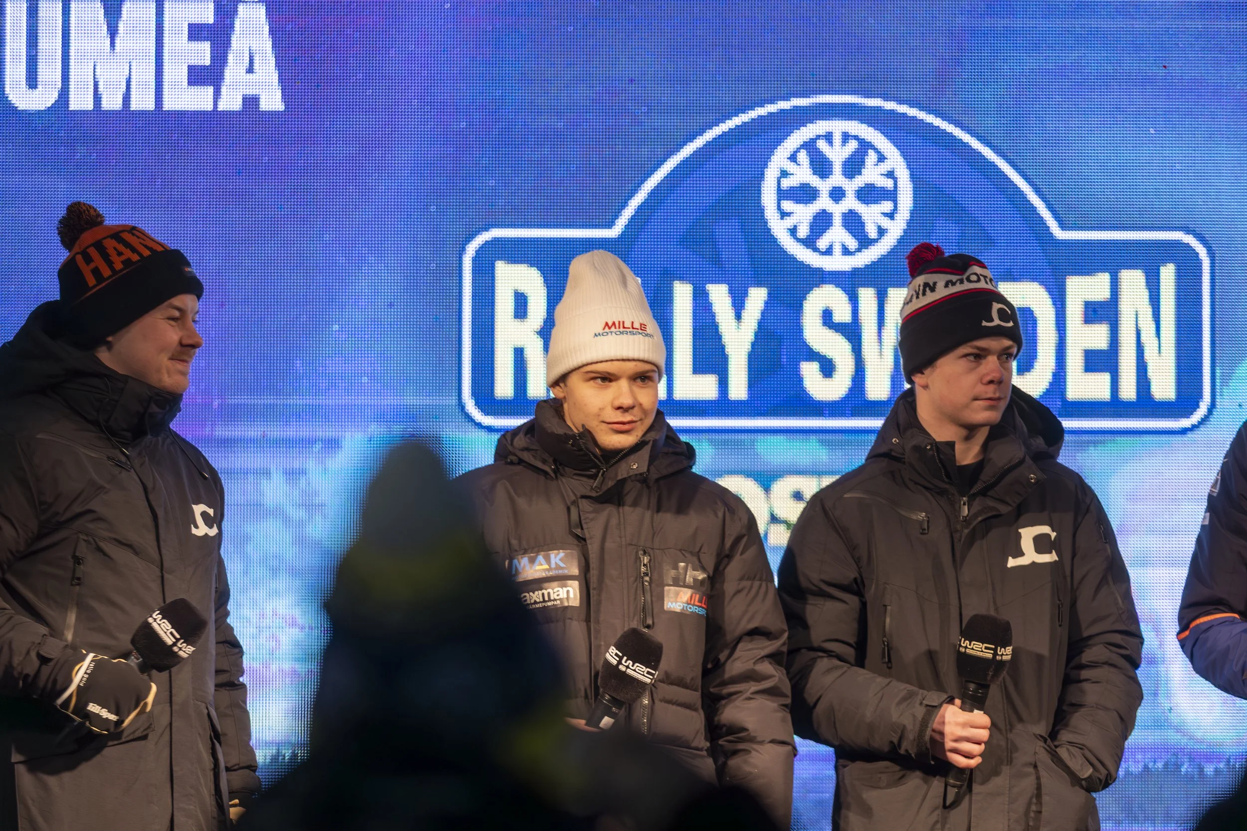 Four men in black winter jackets and hats, holding microphones, standing in front of a blue digital display with logos and text related to rally racing.