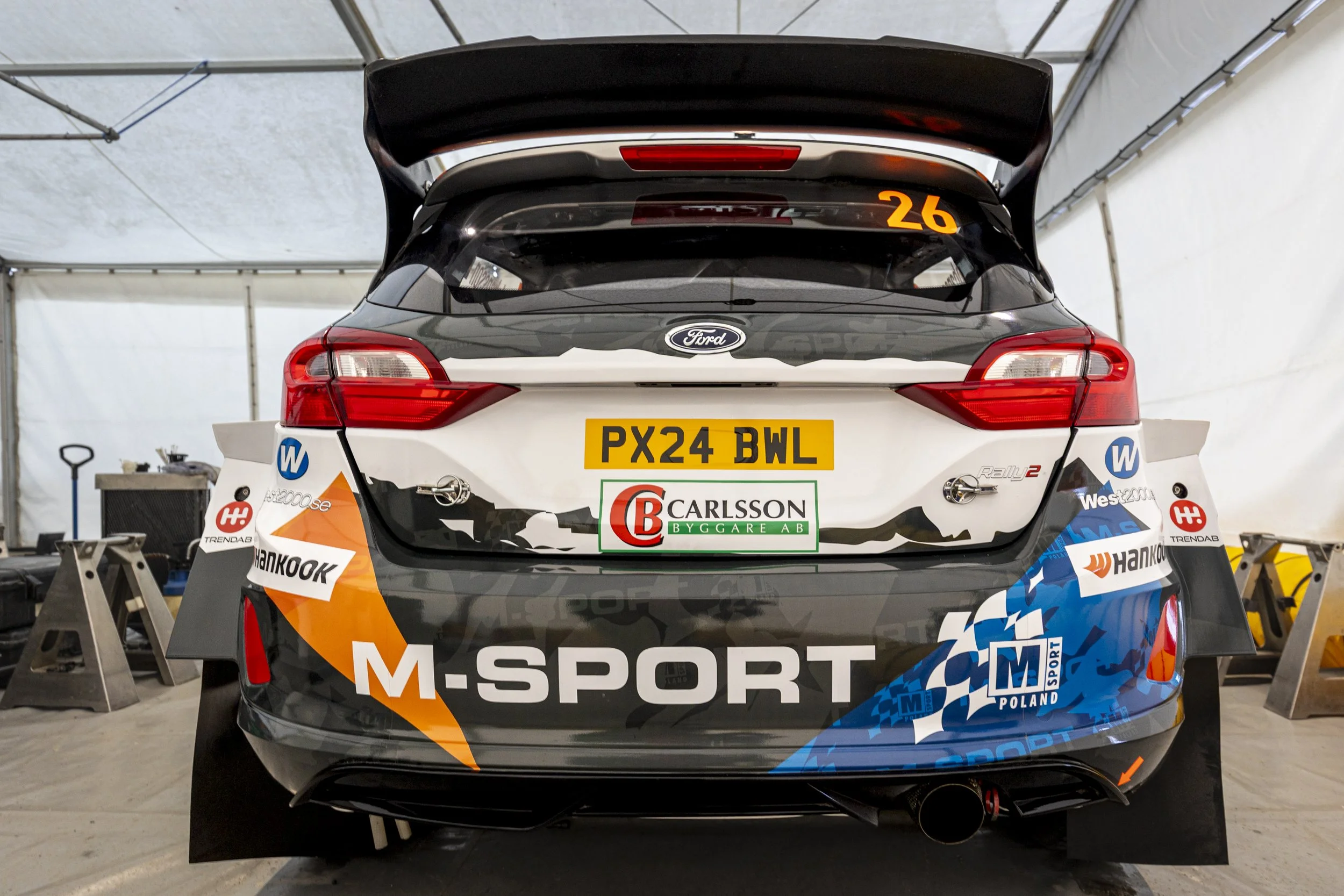 The rear view of a black and white Ford rally car with racing stickers and decals, elevated on a car stand inside a tent with tools and equipment visible in the background.