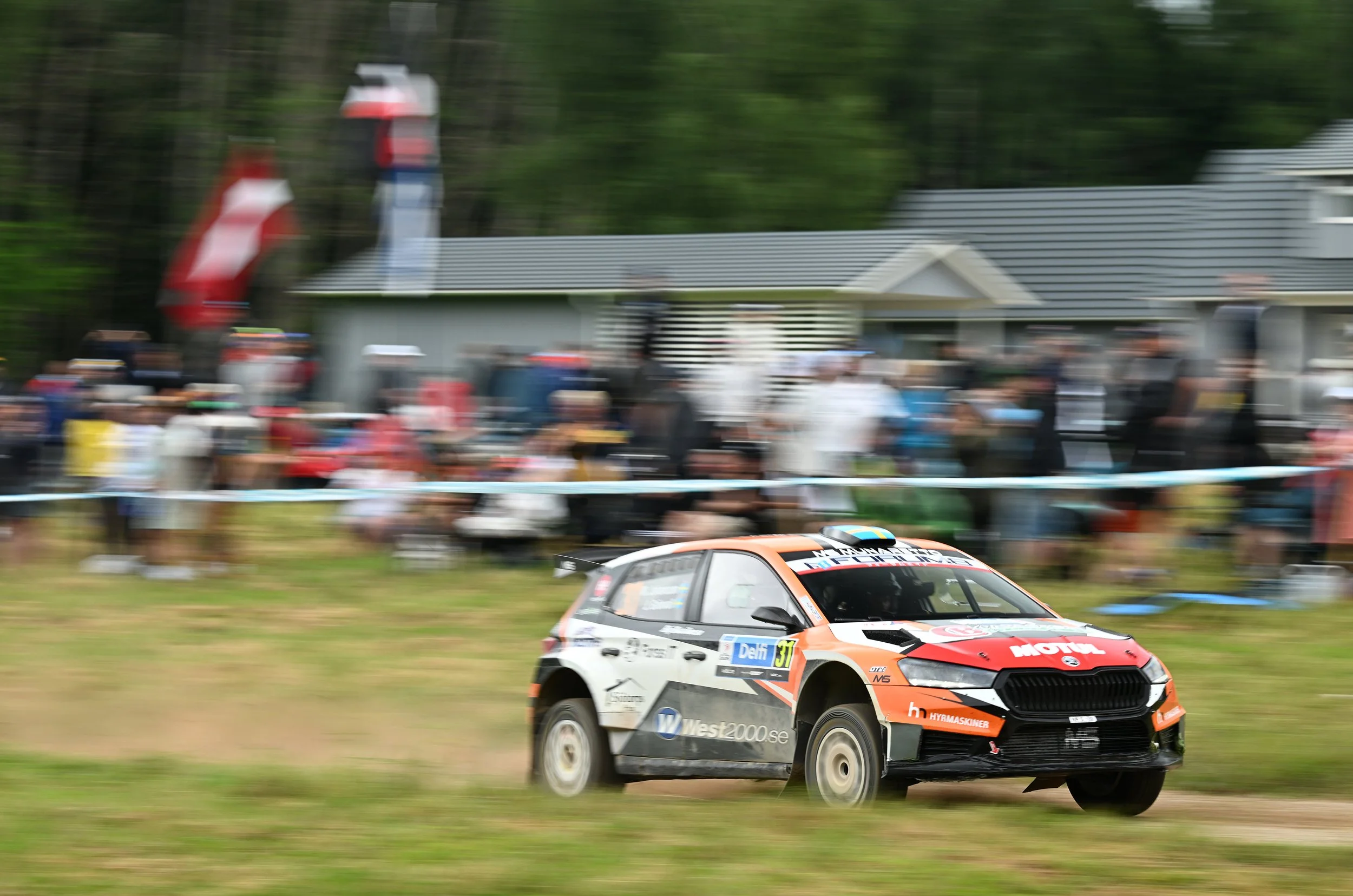 A rally car racing on a dirt track during a motorsport event, with spectators in the background and trees lining the area.
