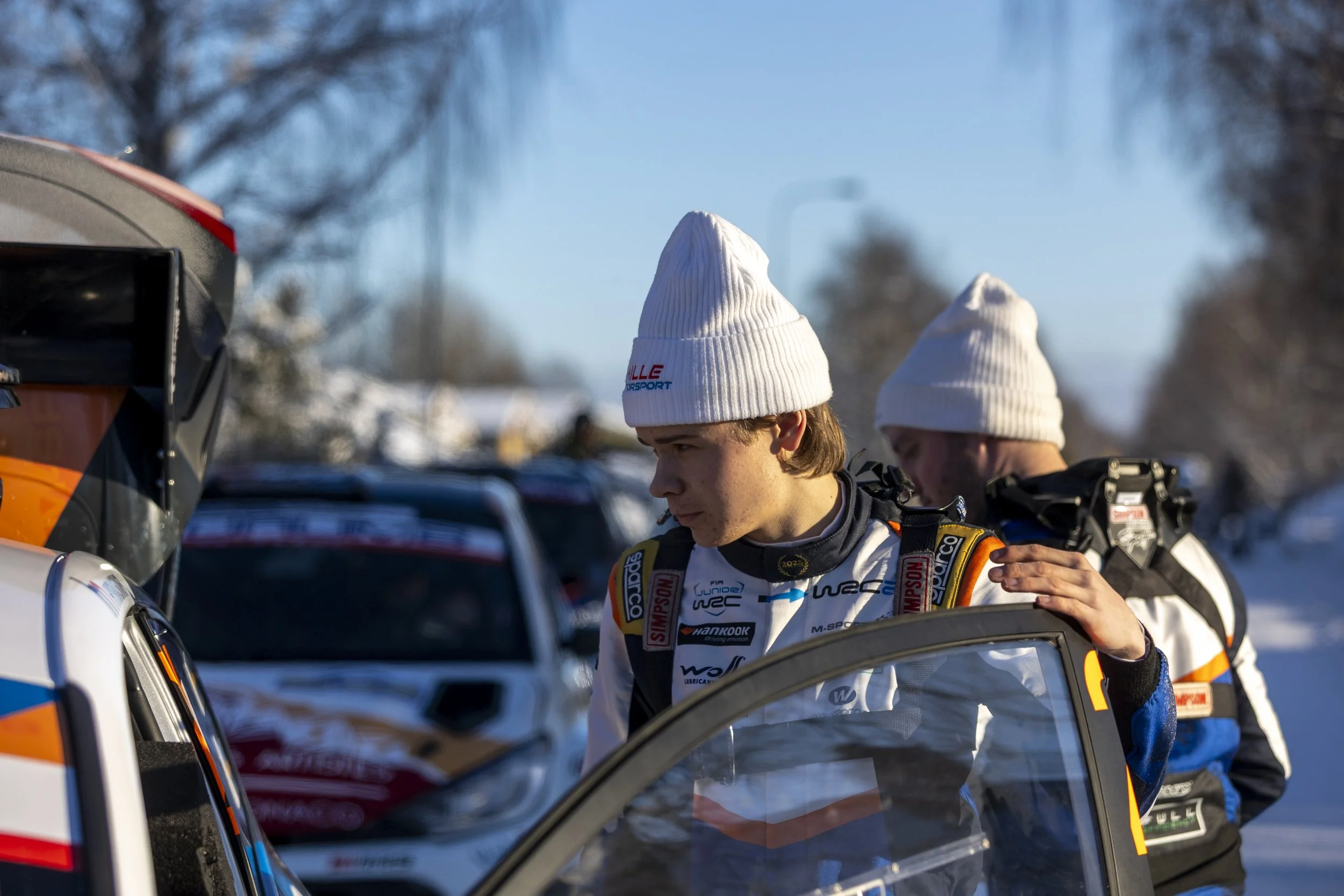 Two young men wearing white beanies and racing suits standing next to race cars, with one opening a car door in a parking lot on a clear day.