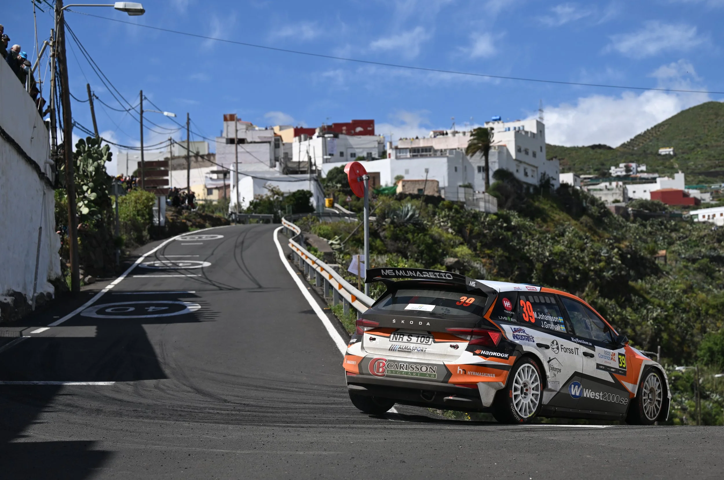 A rally car on a winding mountain road during a race, with a white hillside village in the background and spectators watching from the side.