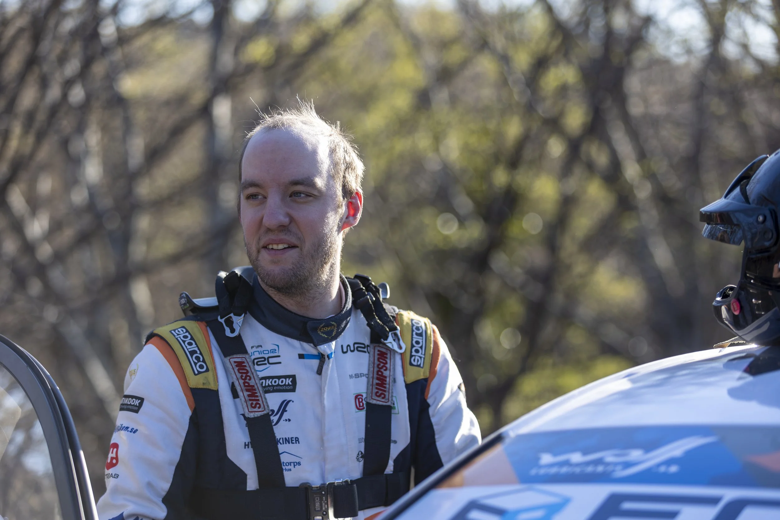 A male rally car driver in a racing suit standing outdoors with trees in the background, wearing a harness, with a racing helmet being handed to him.