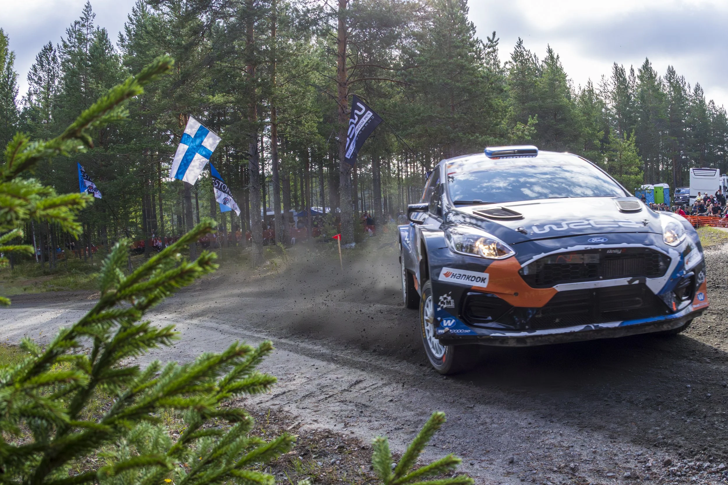 A rally car racing on a dirt track through a forest, kicking up dust with spectators and flags in the background.