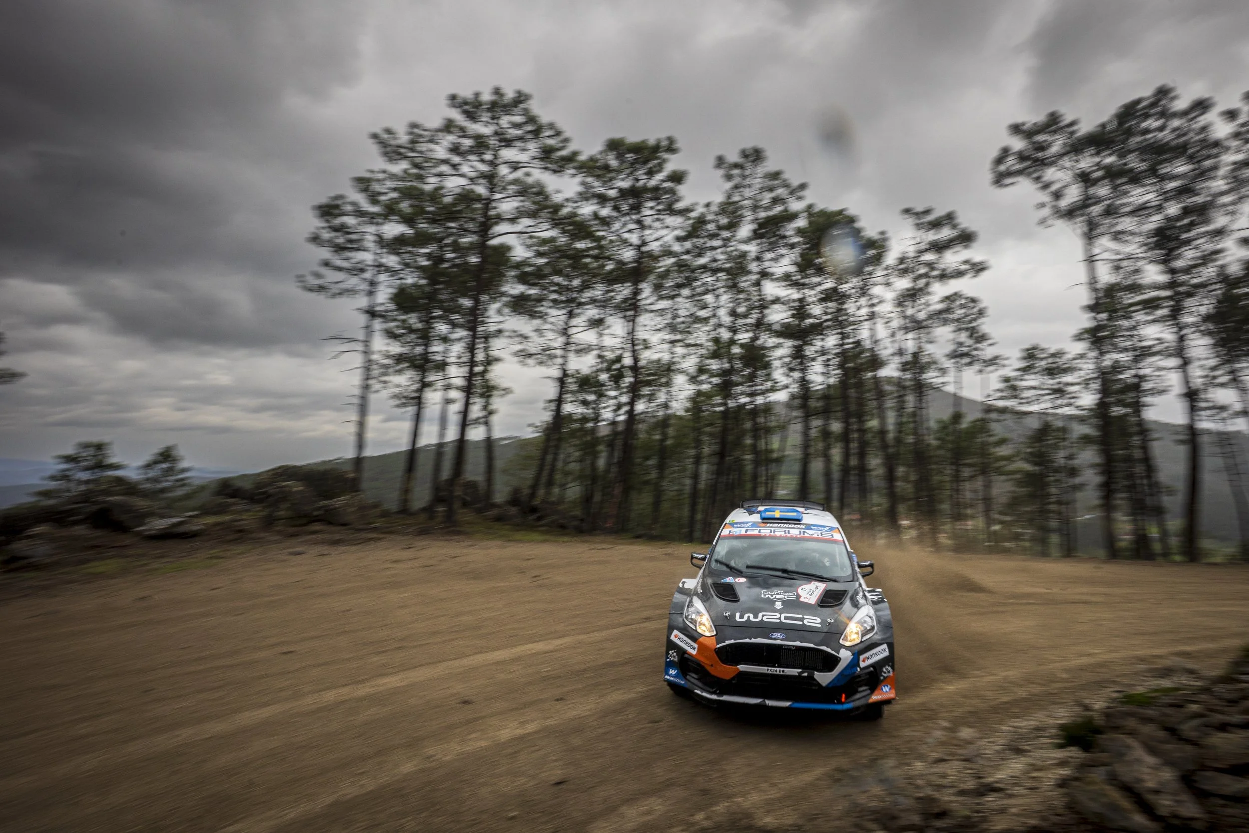 A rally car racing on a dirt road under cloudy skies in a forested area.