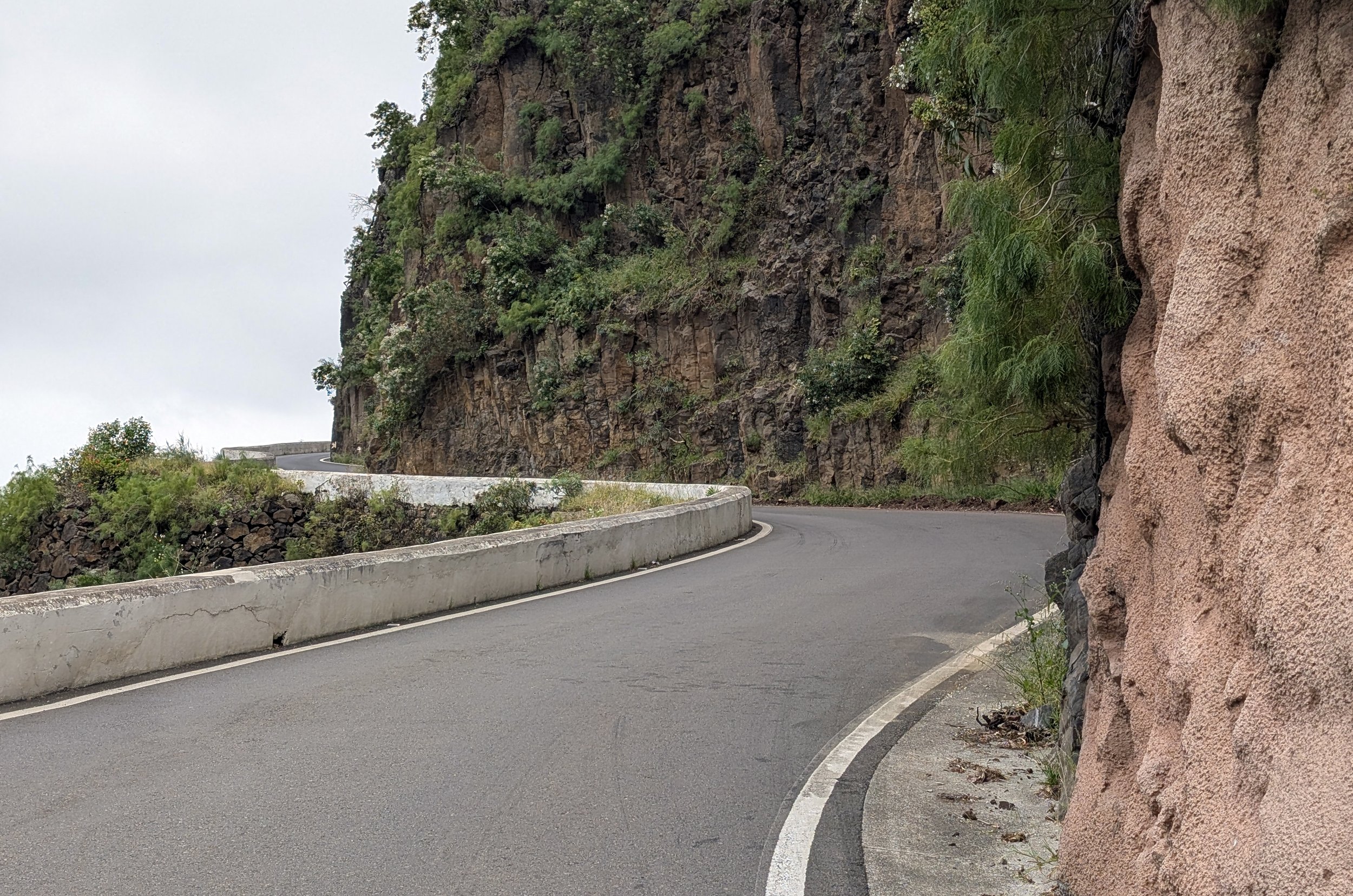 A winding mountain road with a guardrail on the left side and rocks and greenery on the right side, ascending along a rocky hillside under an overcast sky.