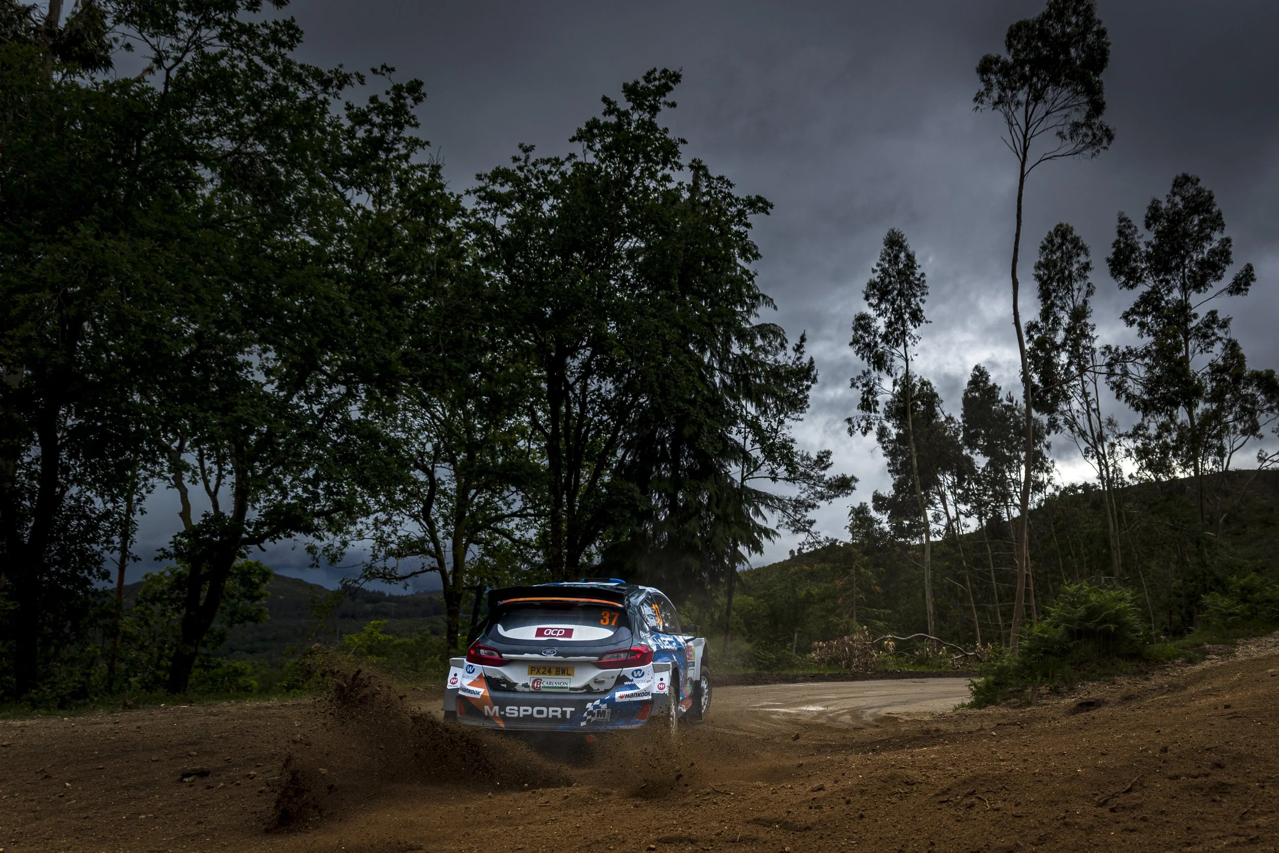 A rally car racing on a dirt road amidst a forest with tall trees and dark, cloudy sky.