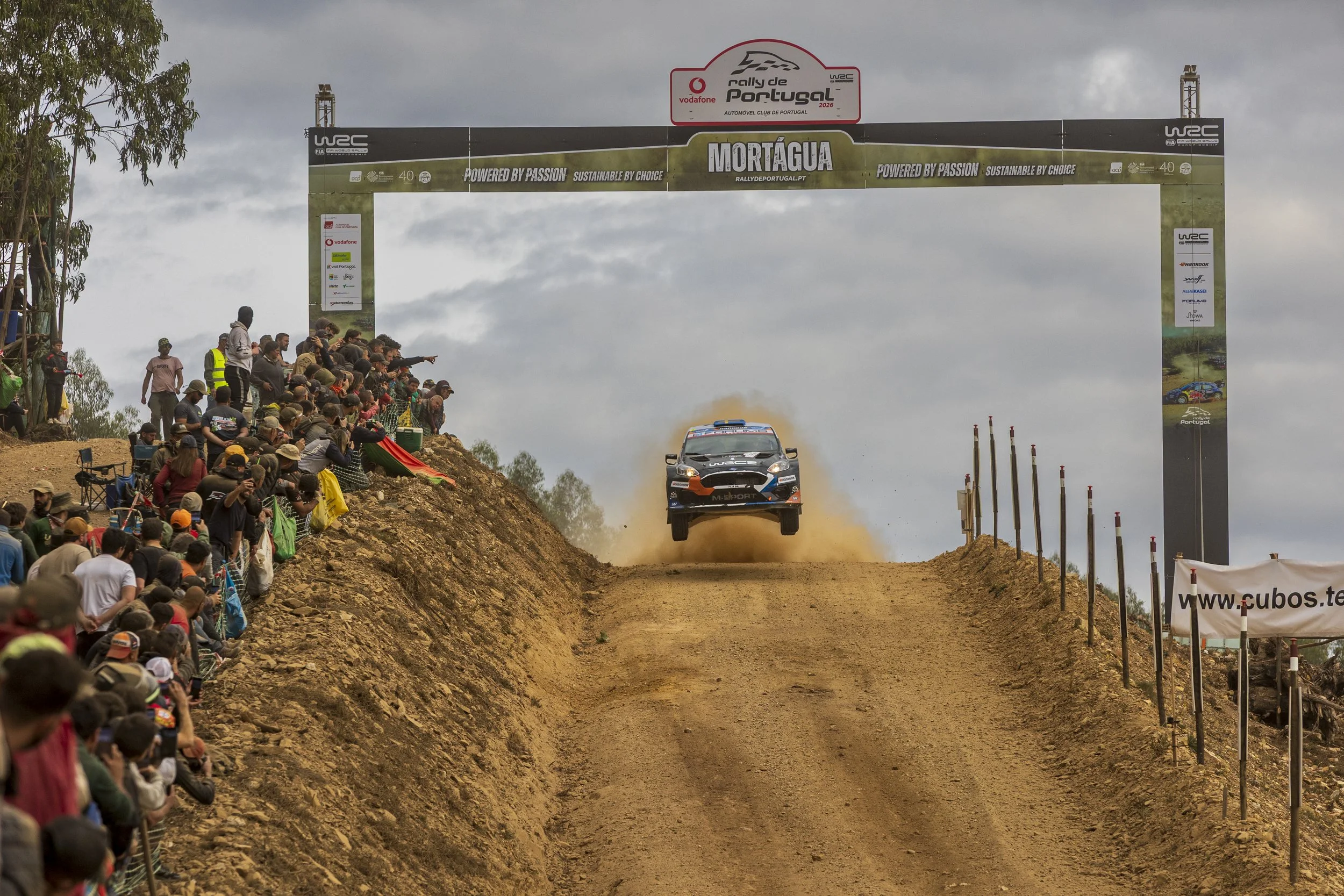 A rally car flying through the air on a dirt track during a race, with spectators watching from both sides.