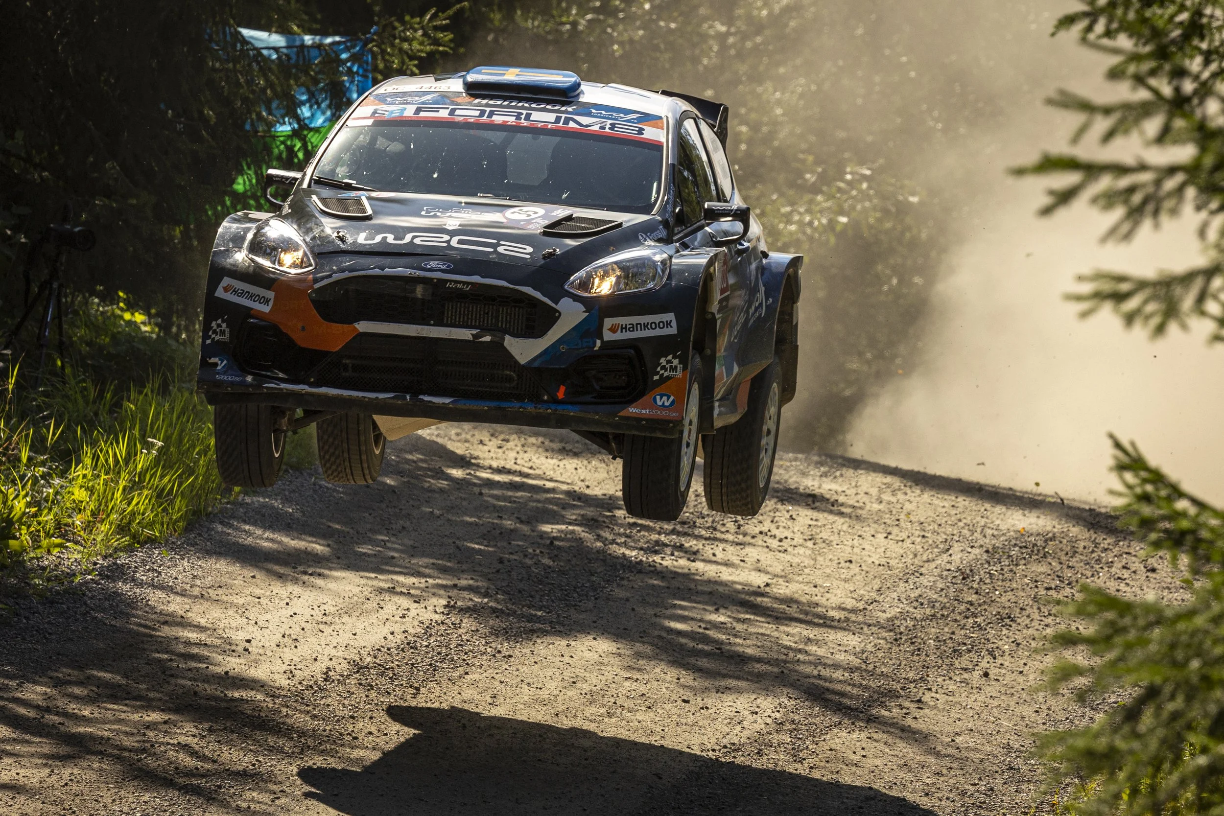 A rally car flying through the air on a dirt road surrounded by trees.