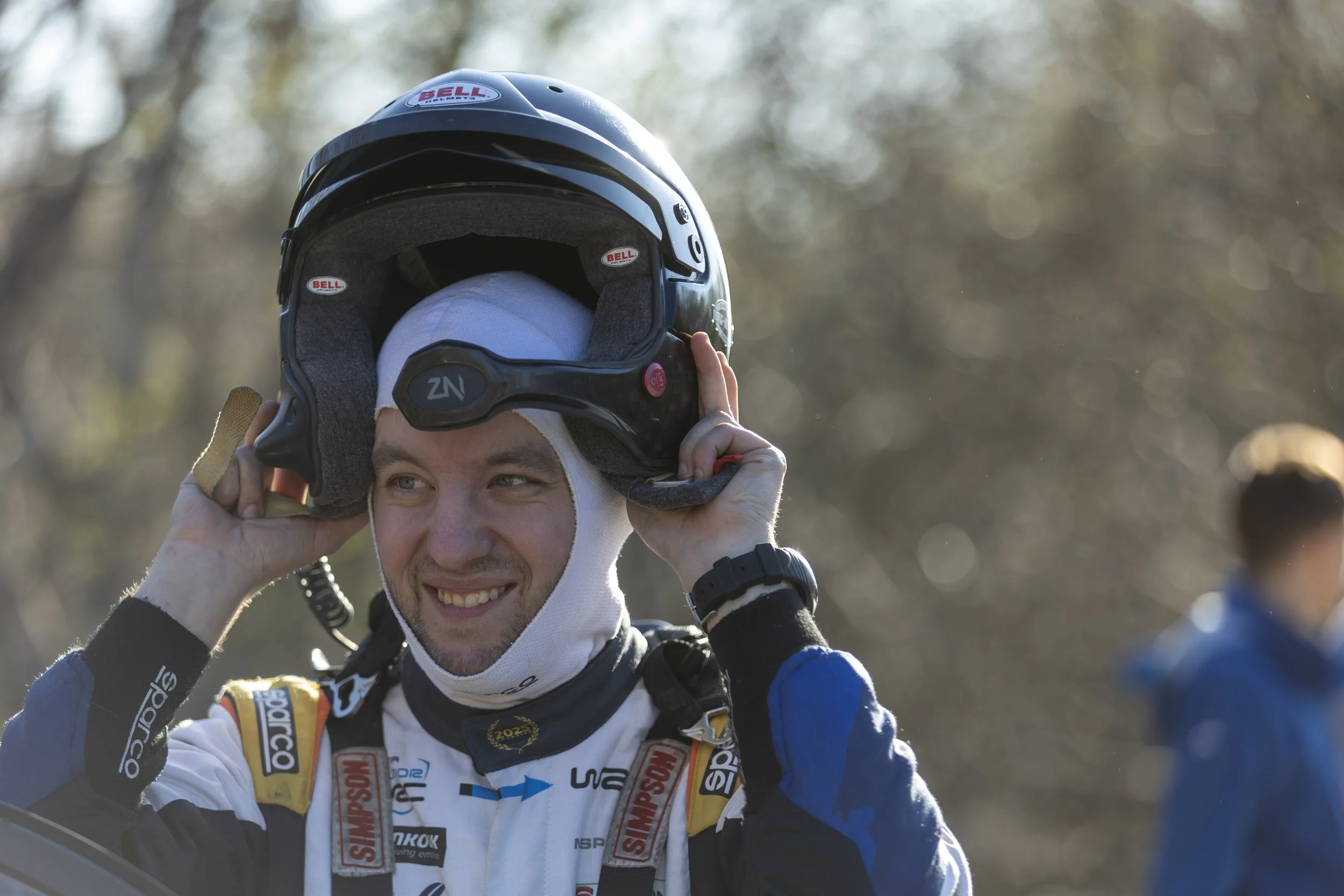 A person is smiling and adjusting a black full-face helmet with a Bell logo, wearing a racing suit with sponsor patches, outdoors with a blurred background of trees and other people.