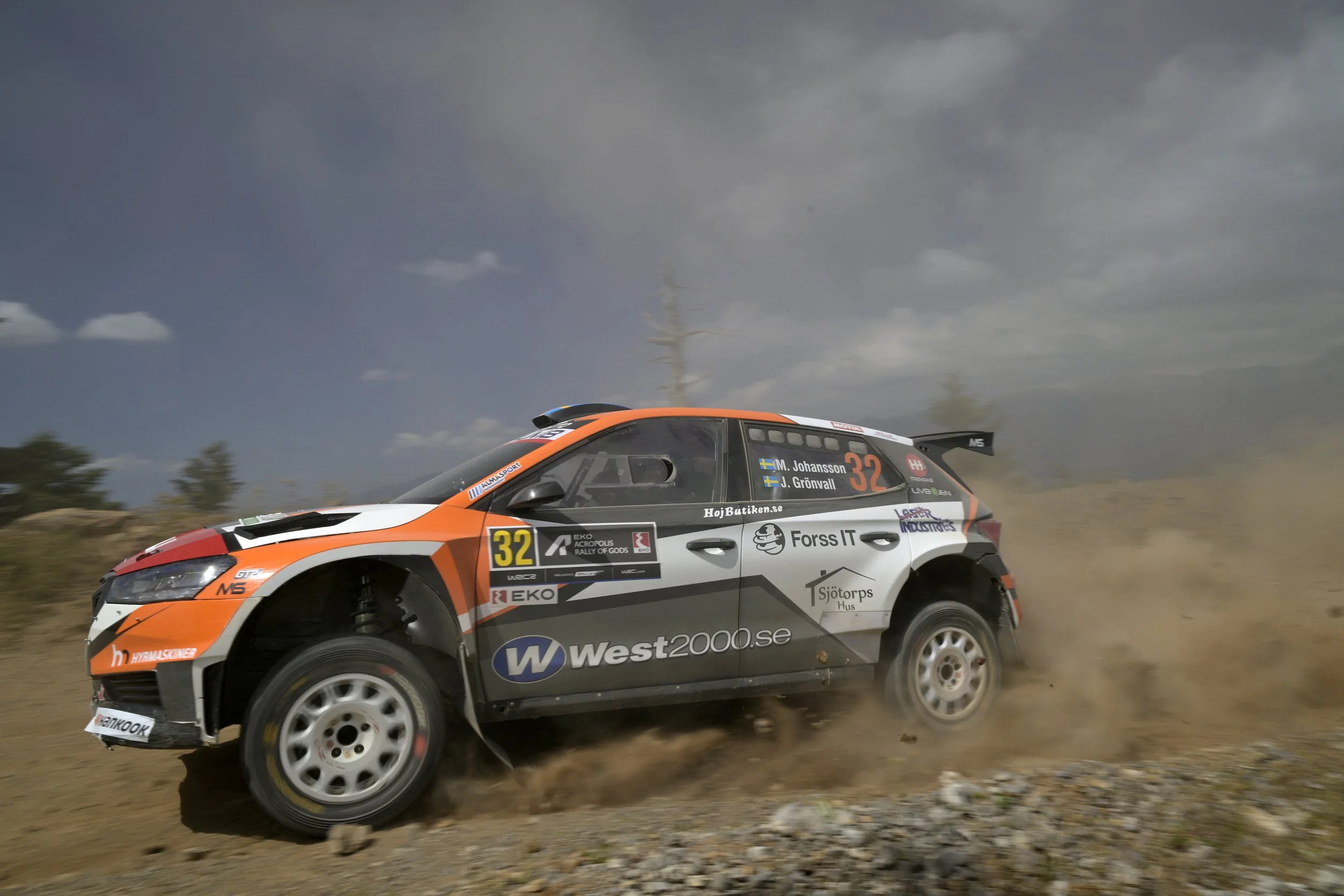 A rally car speeding on a dirt road, kicking up dust, with a cloudy sky in the background.