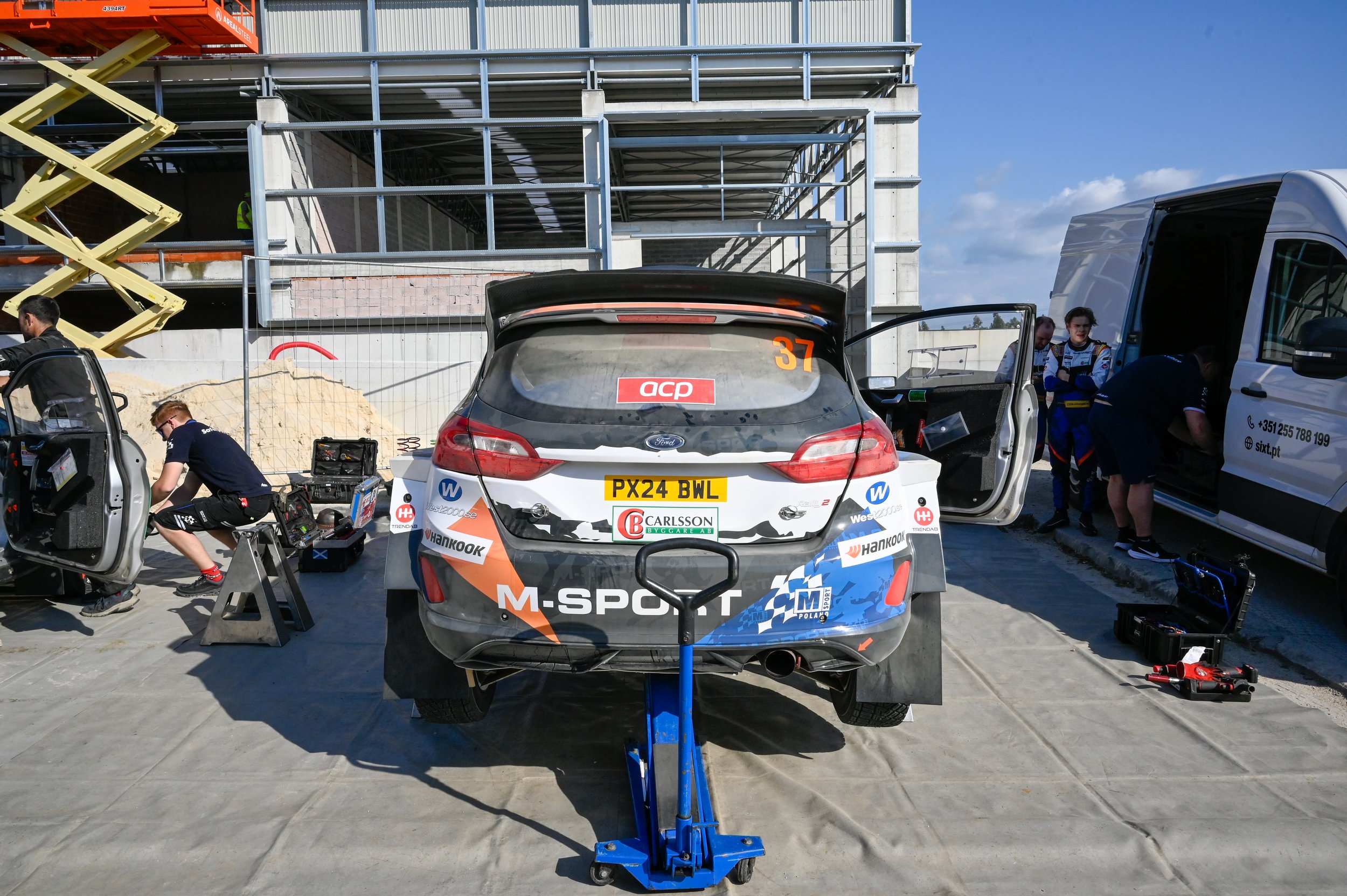 Race car being repaired in a pit area with mechanics working around it, scaffoldings, and construction site elements in the background.