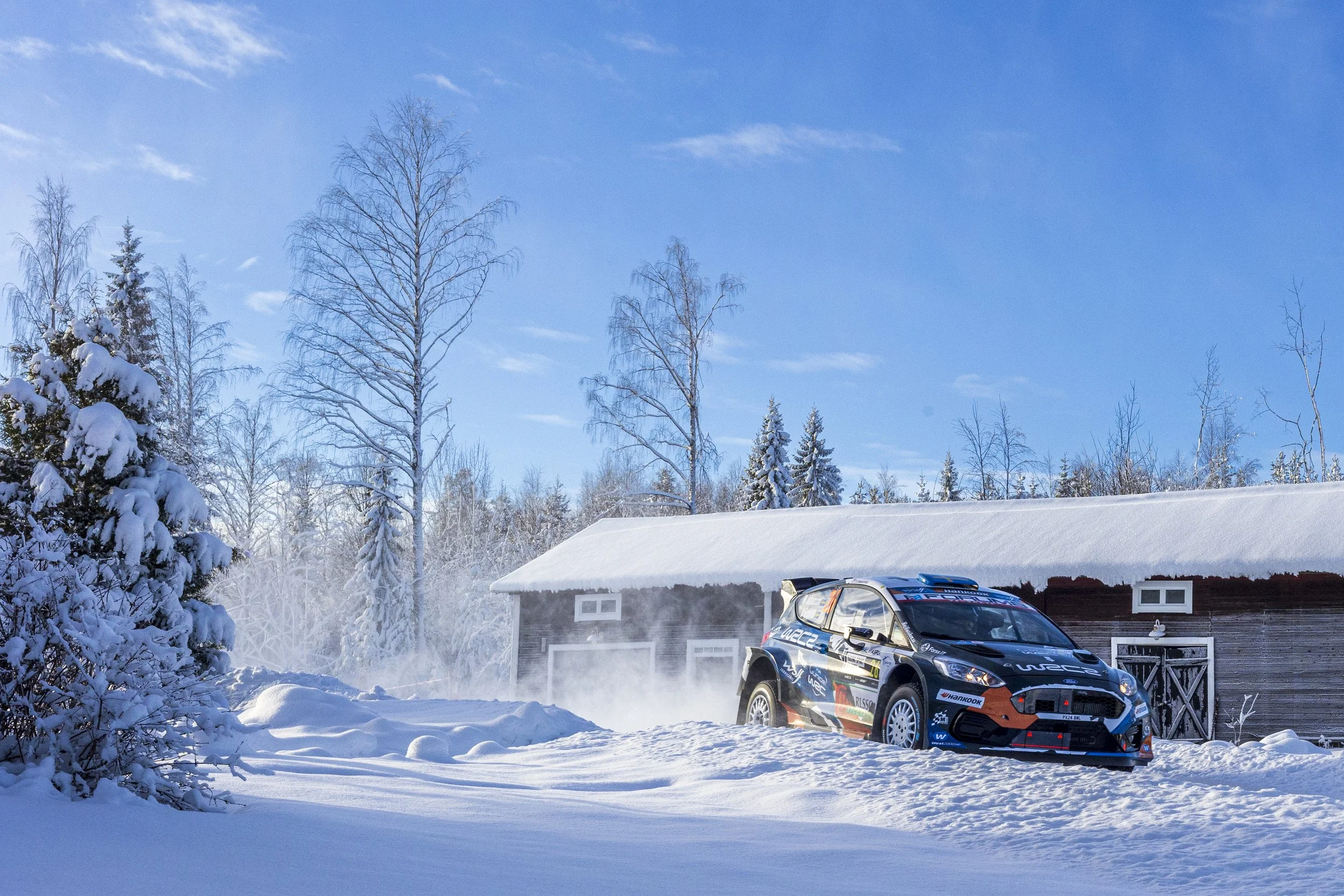 A snow-covered landscape with trees and a building, a rally car on the snow, and a blue sky with some clouds.