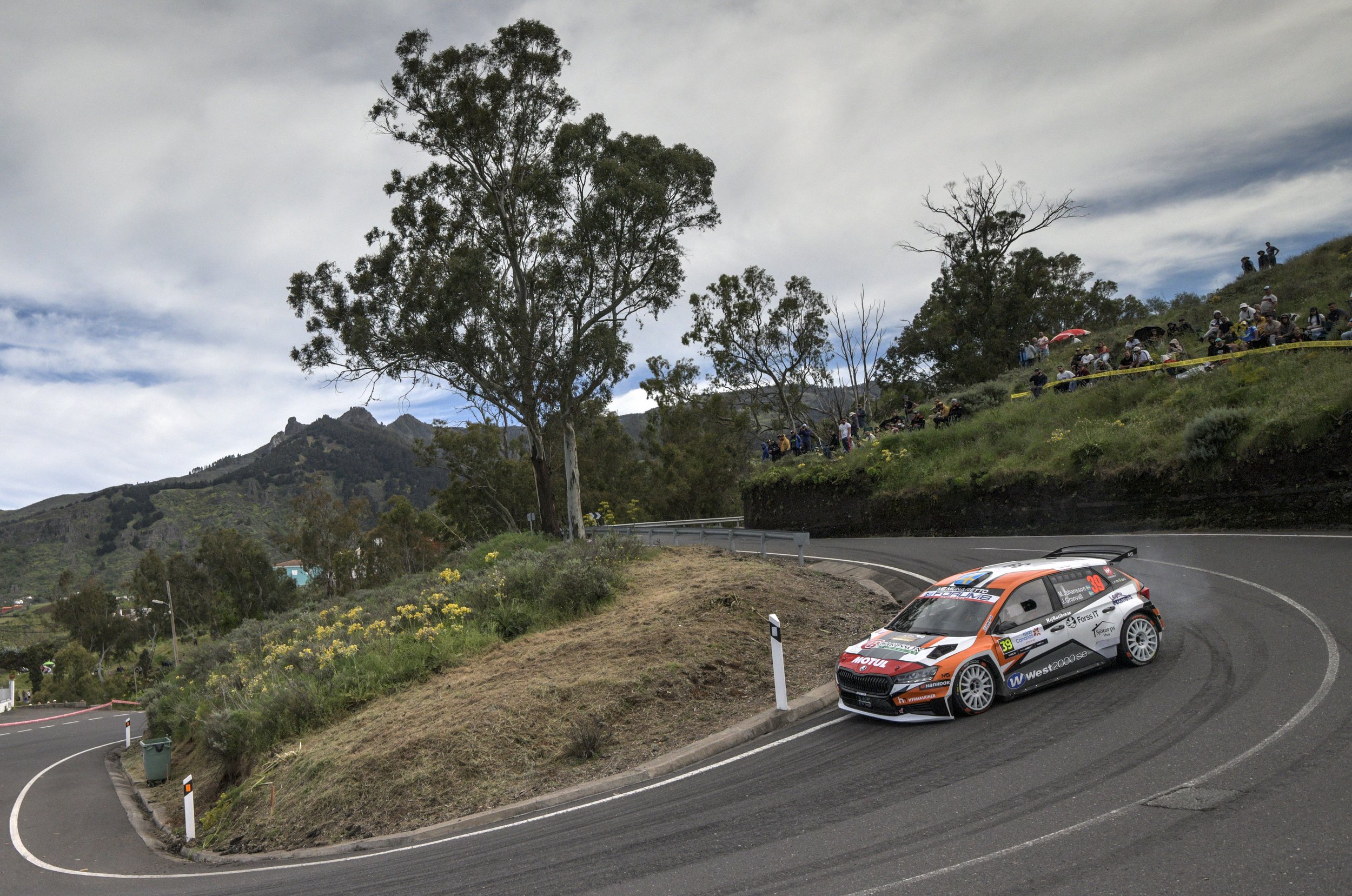 A rally car is navigating a sharp turn on a winding mountain road, with spectators watching from the hillside in the background.