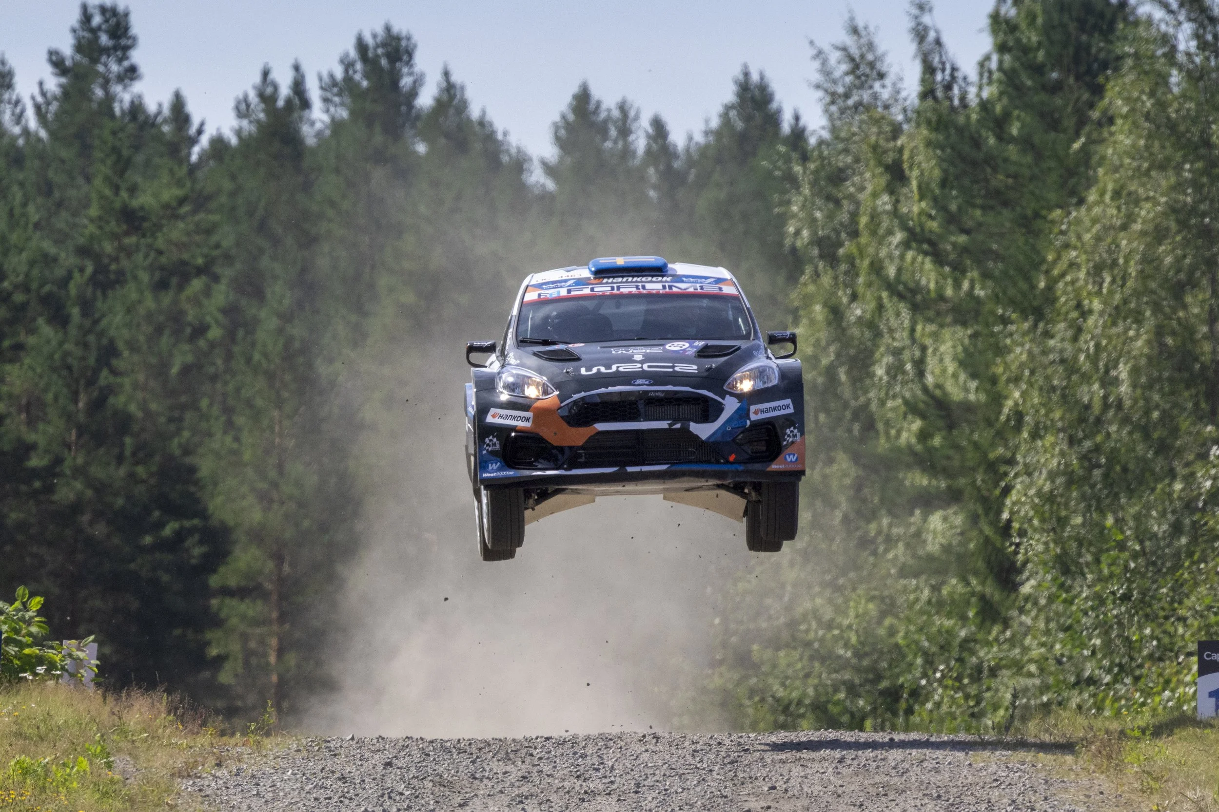 A rally car is airborne on a dirt road surrounded by trees during a race.