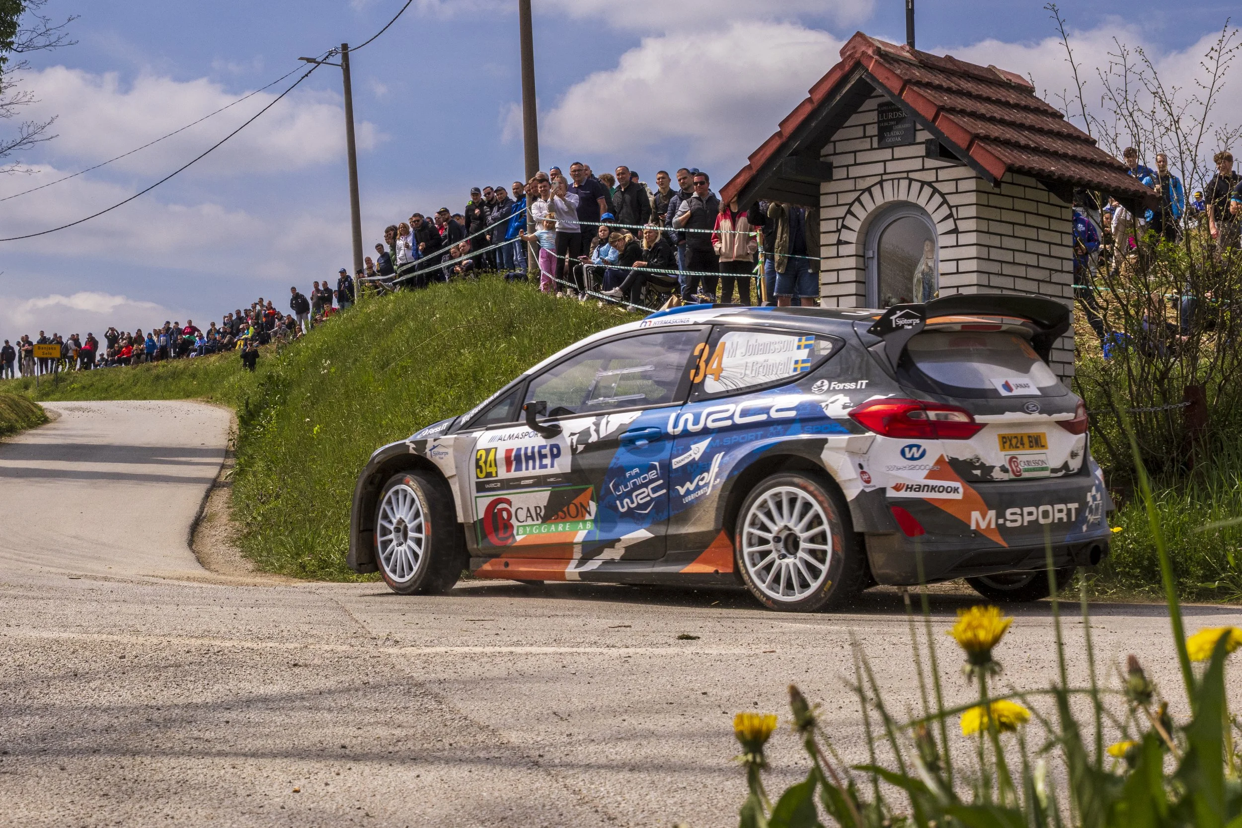 A rally car on a dirt road, with a crowd of people watching from the hillside and nearby structures, under a partly cloudy sky.