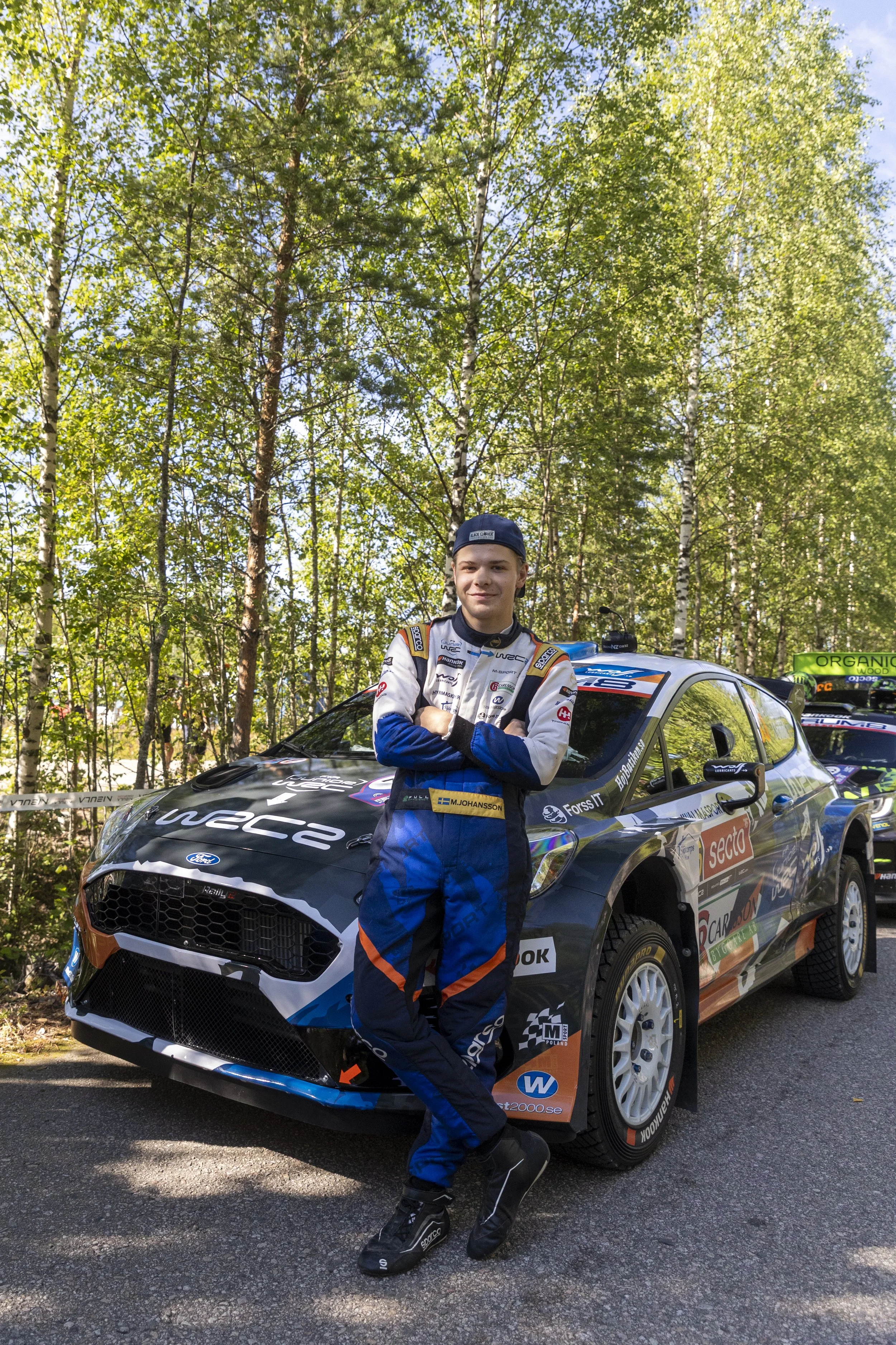 A young rally car driver in racing suit and cap, standing in front of a black rally car with sponsor stickers, in a forest setting with trees and sunlight.
