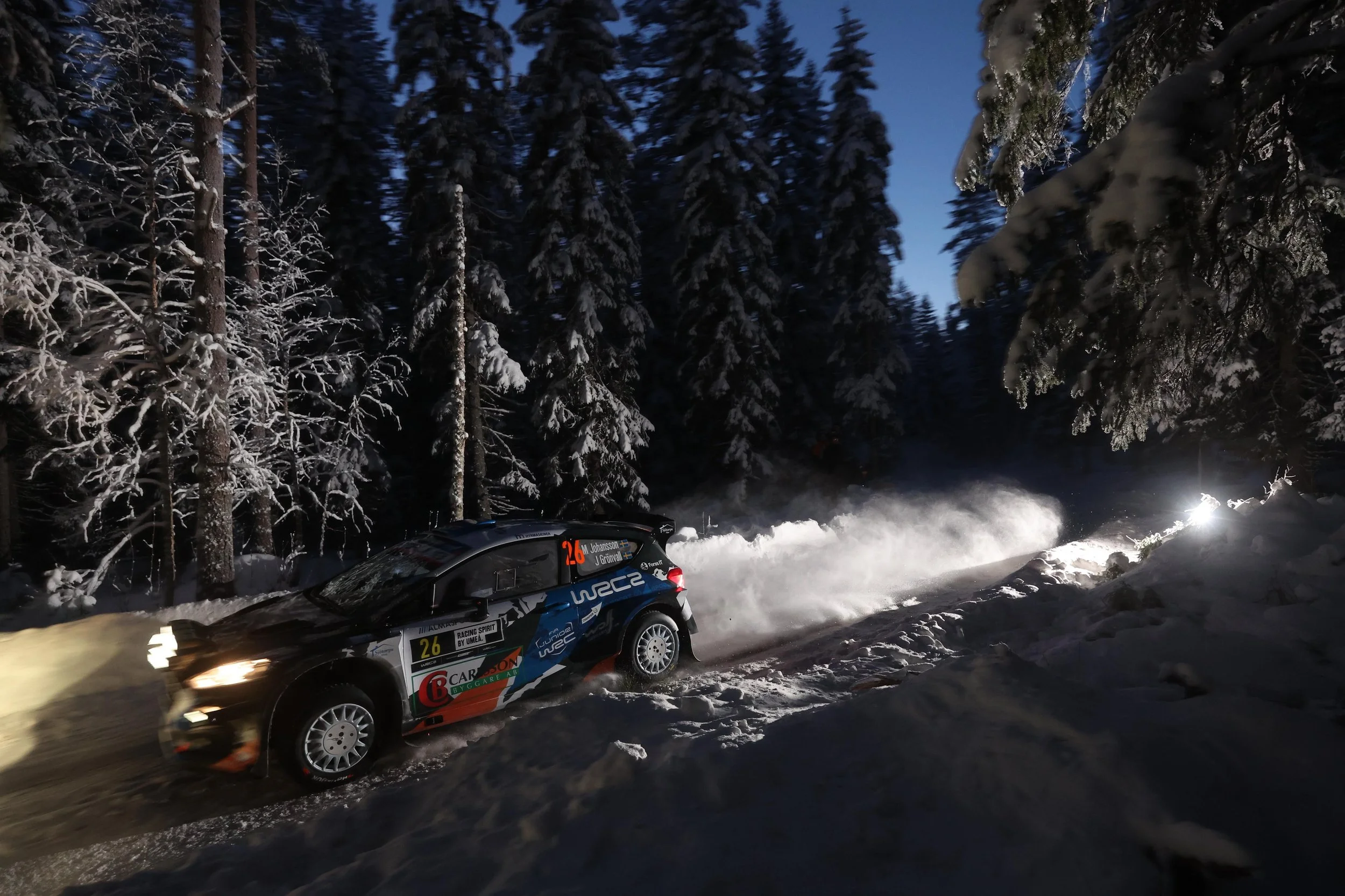A rally car speeding through a snowy forest trail at night, kicking up snow behind it. The car has sponsor stickers and a racing number, with tall snow-covered trees on either side.