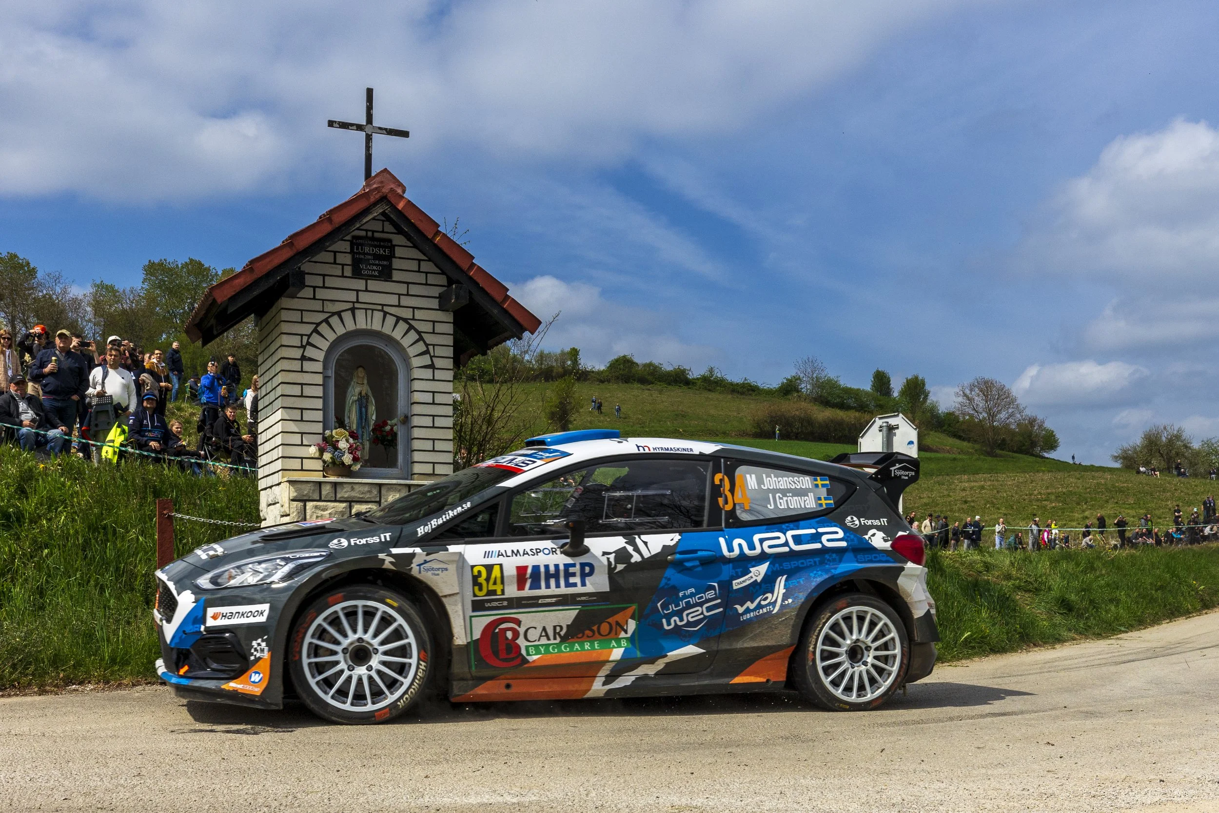 A rally car with the number 34 racing past a small shrine and a crowd of spectators on a grassy hill under a partly cloudy sky.
