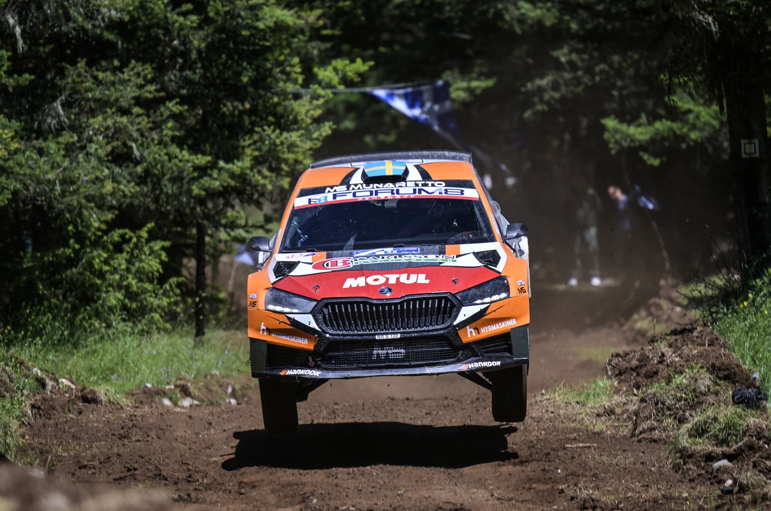 An orange rally car is racing on a dirt road through a lush green forest, with dust and dirt kicked up behind it.