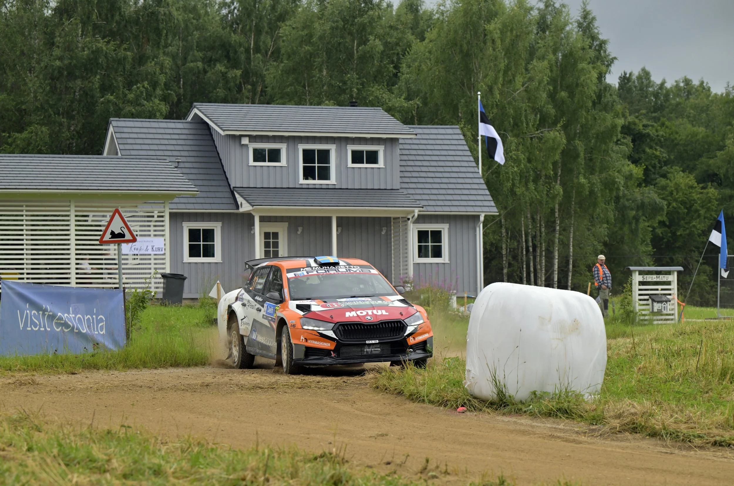 A rally car speeding on a dirt track with a house and trees in the background, flags flying, and a person standing nearby.