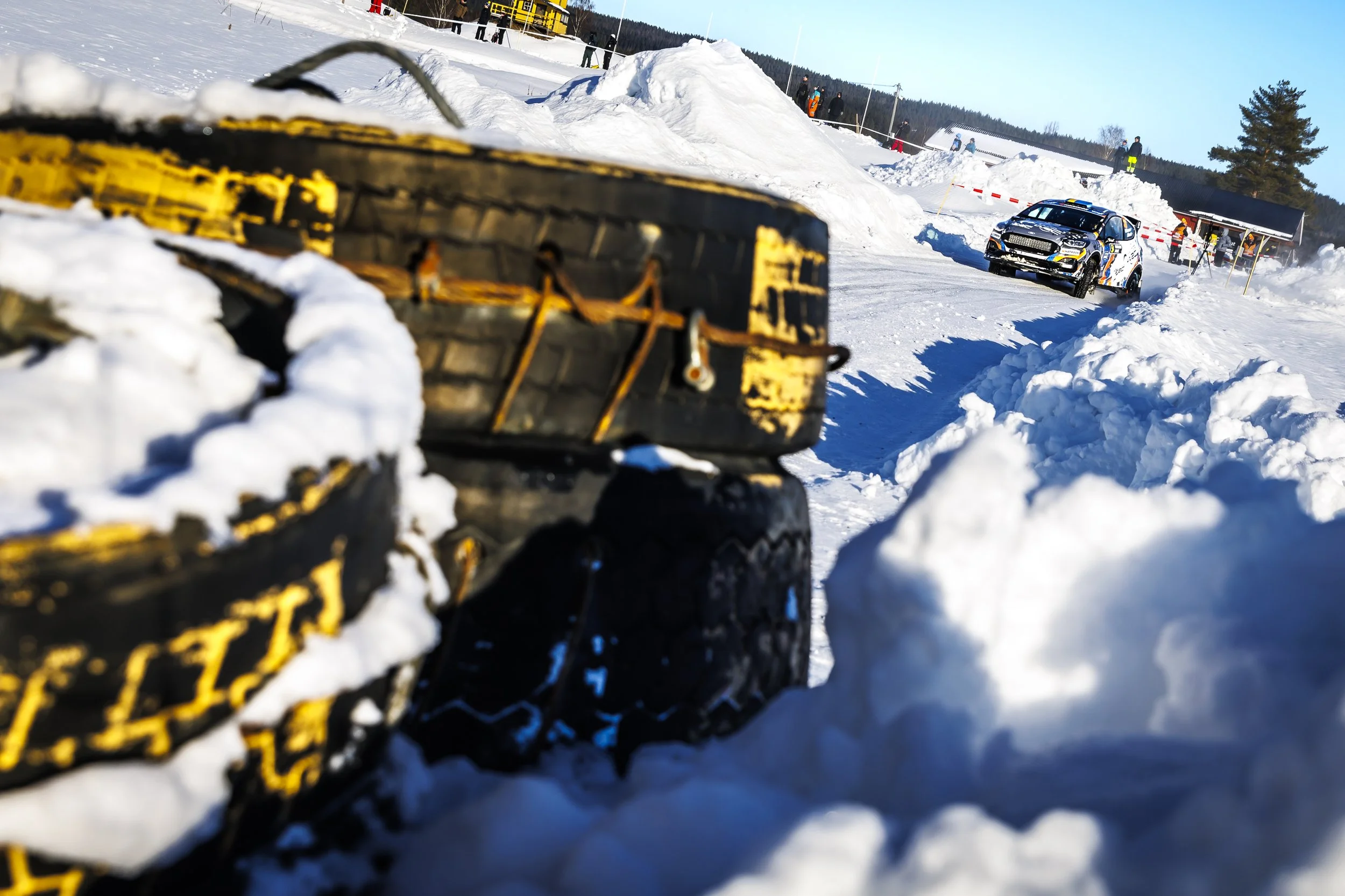 Snow-covered landscape with a rally car driving on a snowy road, spectators in the background, and snowshoes in the foreground.