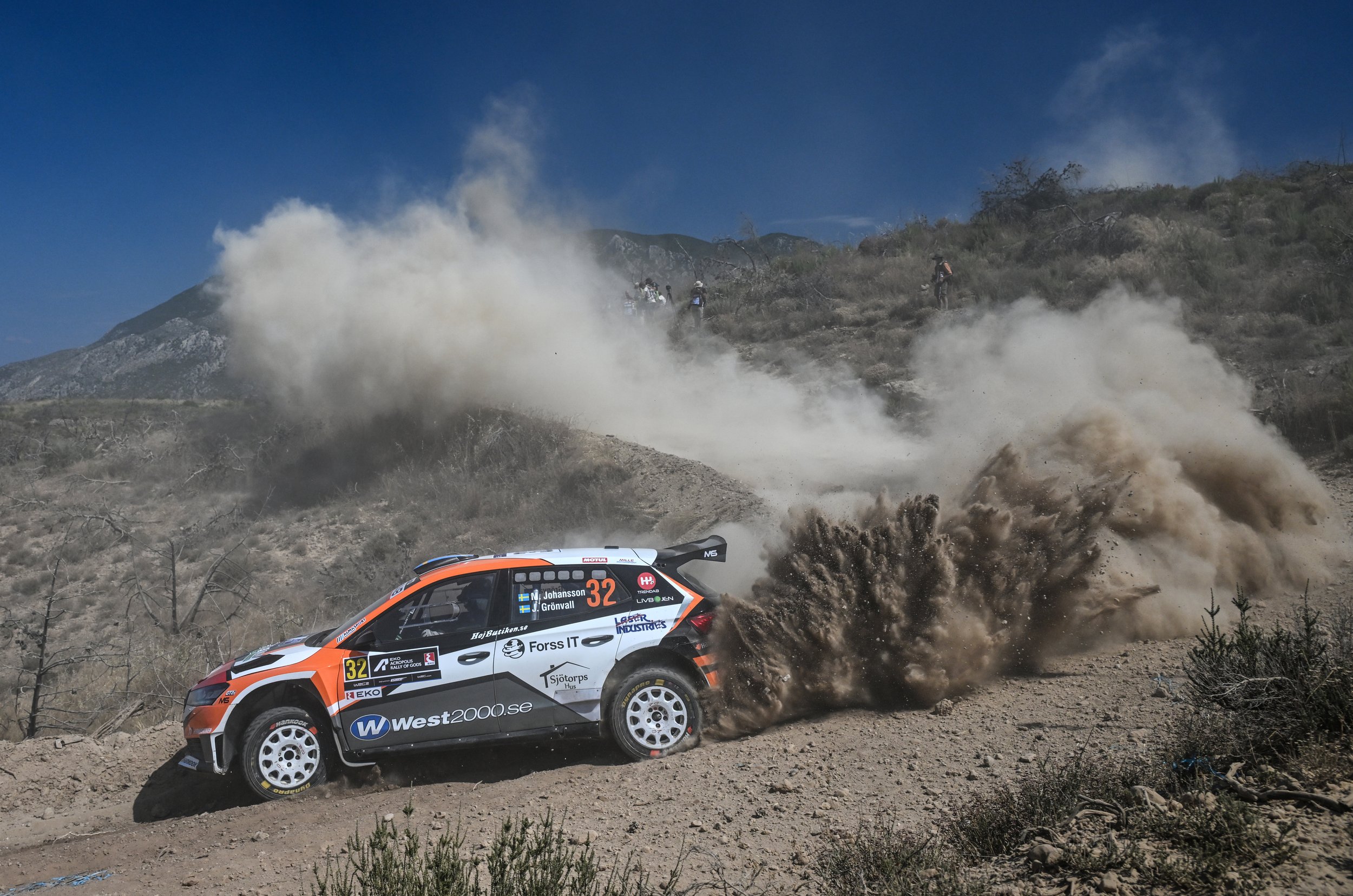 A rally car speeding on a dirt road, kicking up dust and debris, during a race in a rugged outdoor setting.
