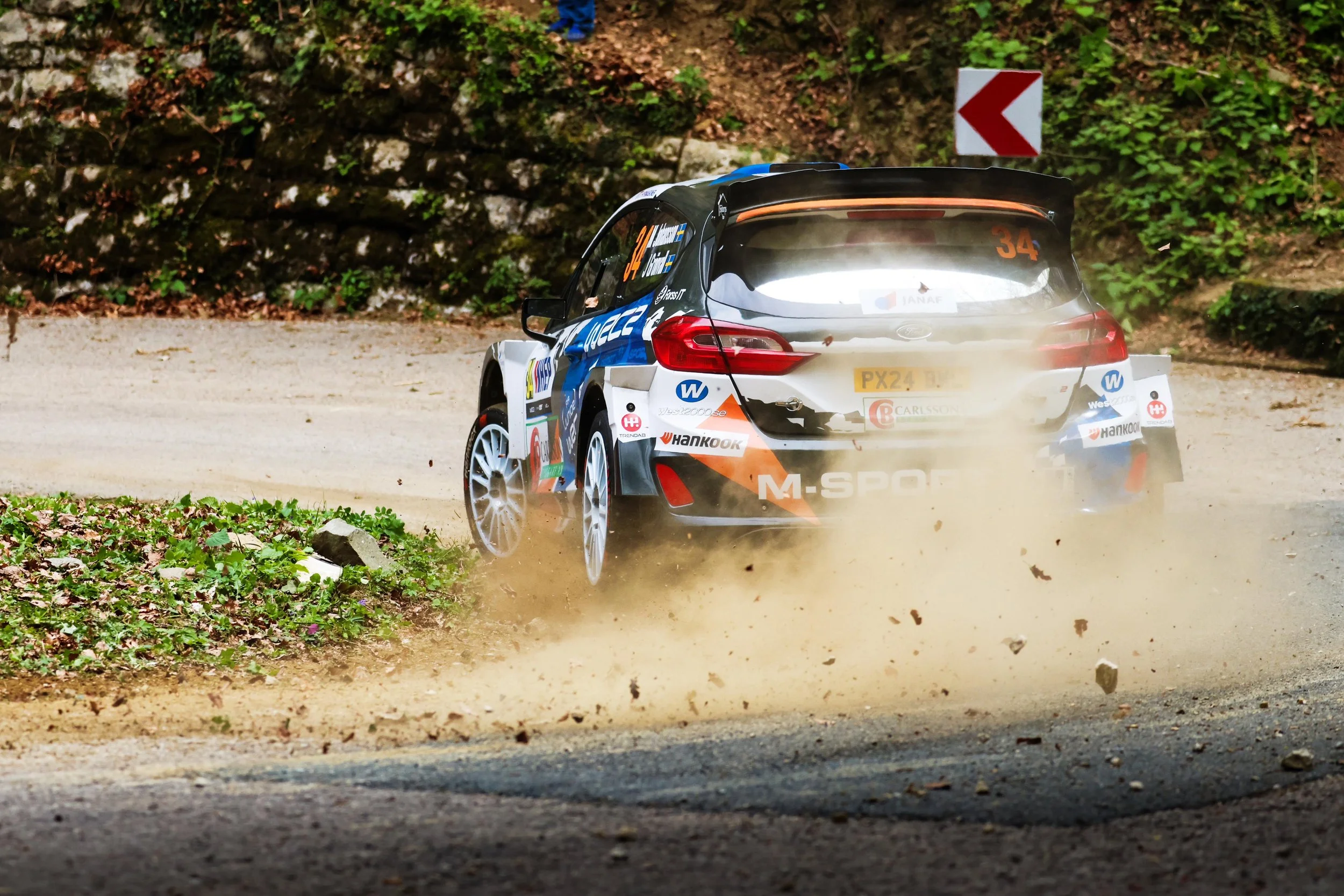 Rally car speeding down a dirt road, kicking up dust, with a rocky and green forested background, and a right turn sign visible.