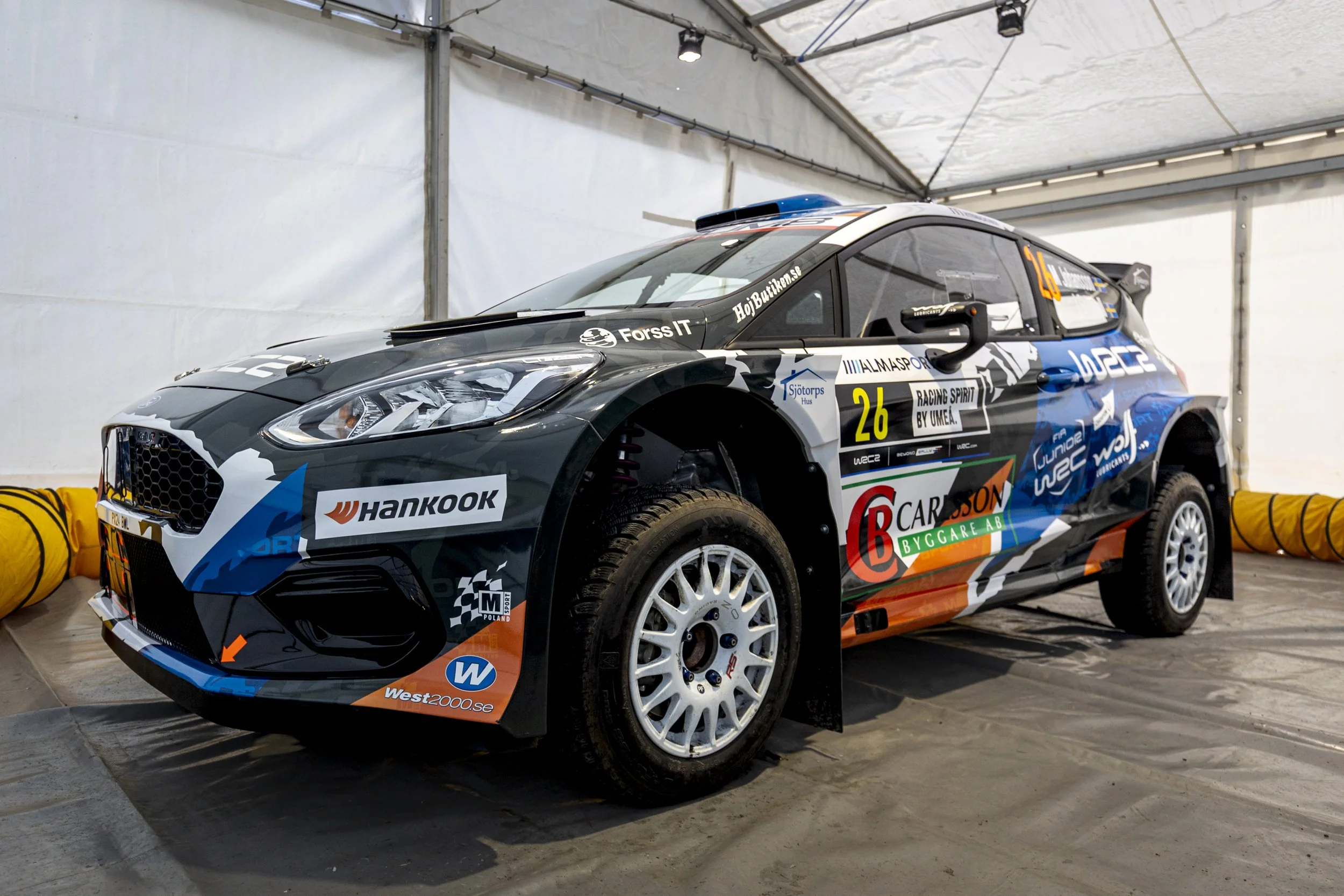 A race car inside a white tent, with various sponsor logos and the number 26 on its side.