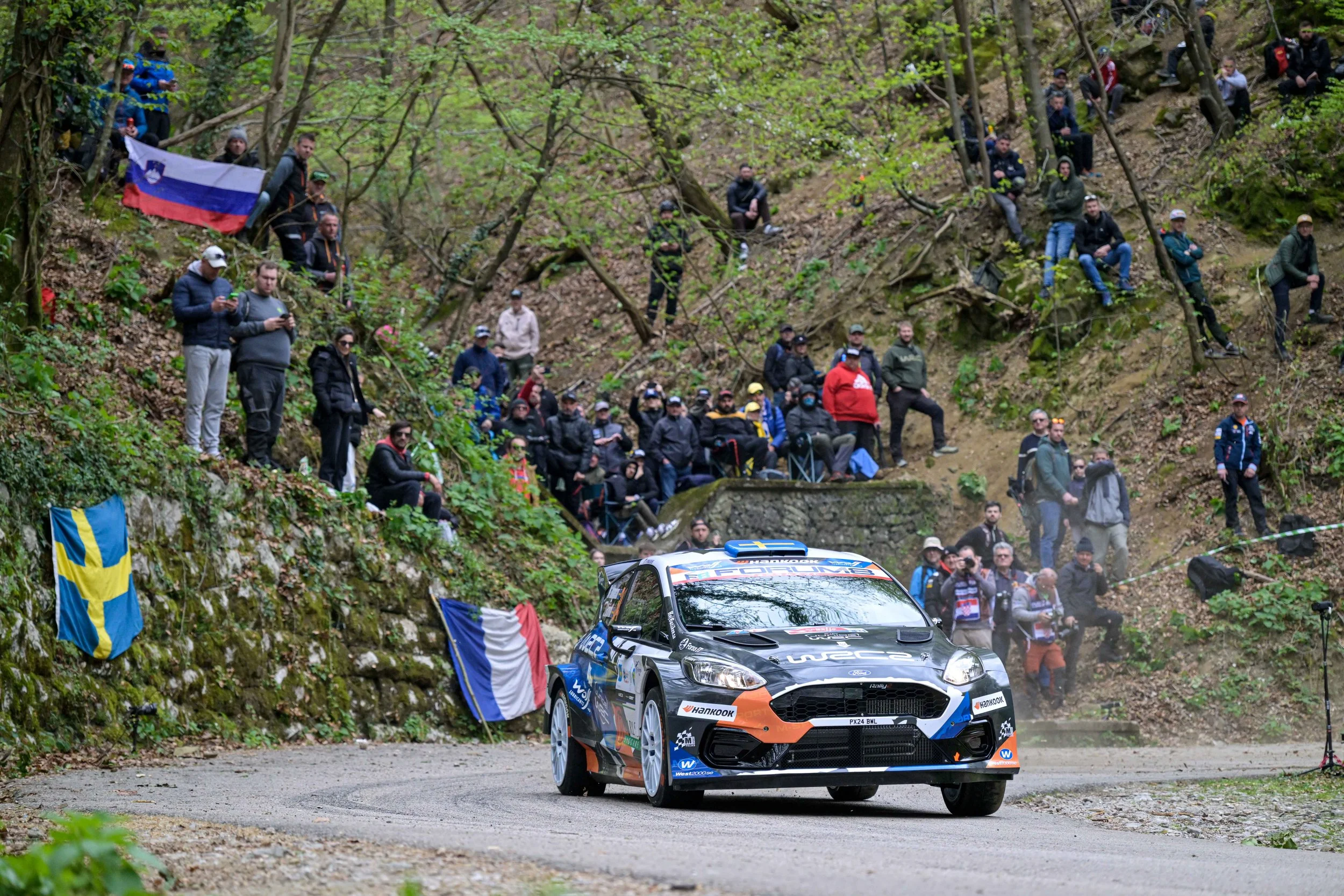 A rally car driving on a dirt road during a race, with spectators watching from a wooded hillside decorated with flags.