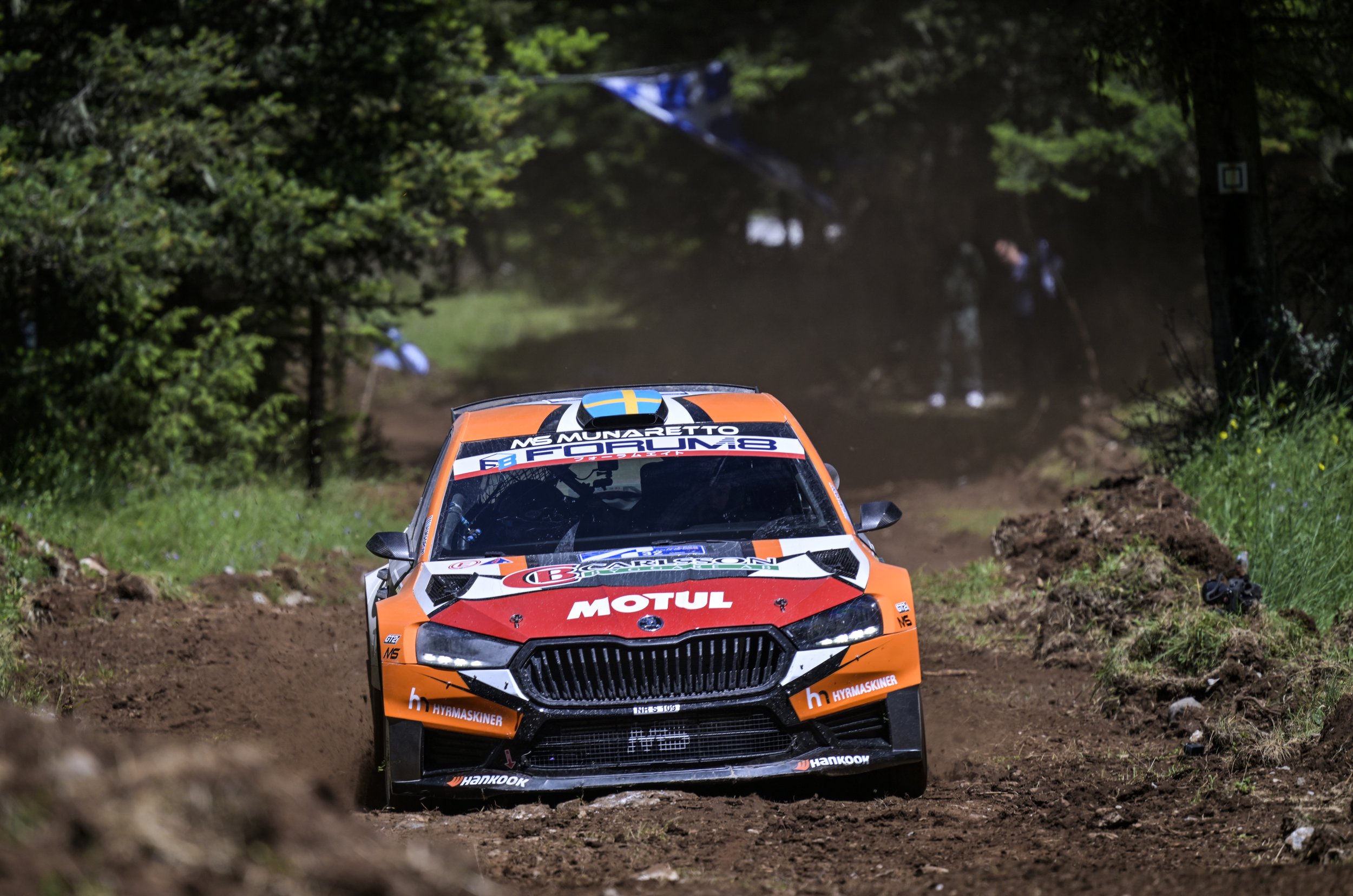 An orange rally car speeding along a dirt road through a forest with trees and foliage on both sides.