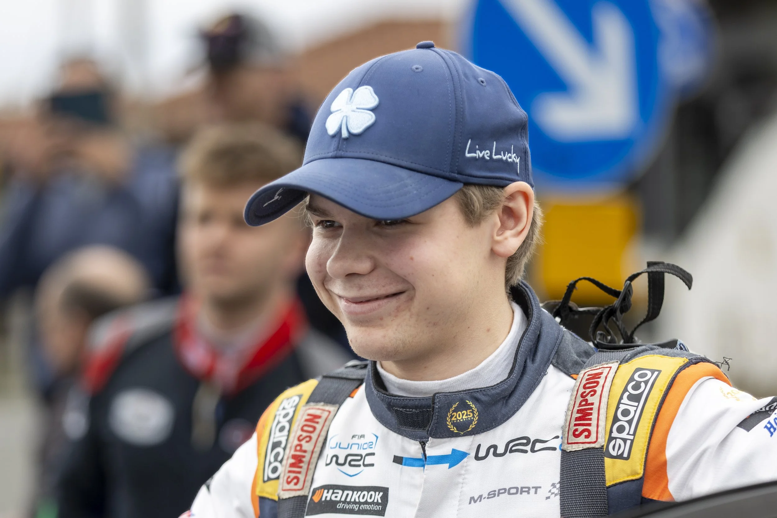 A young race car driver in a racing suit and a blue cap with a four-leaf clover and the phrase 'Live Lucky' embroidered on it, smiling and standing in a crowded setting.