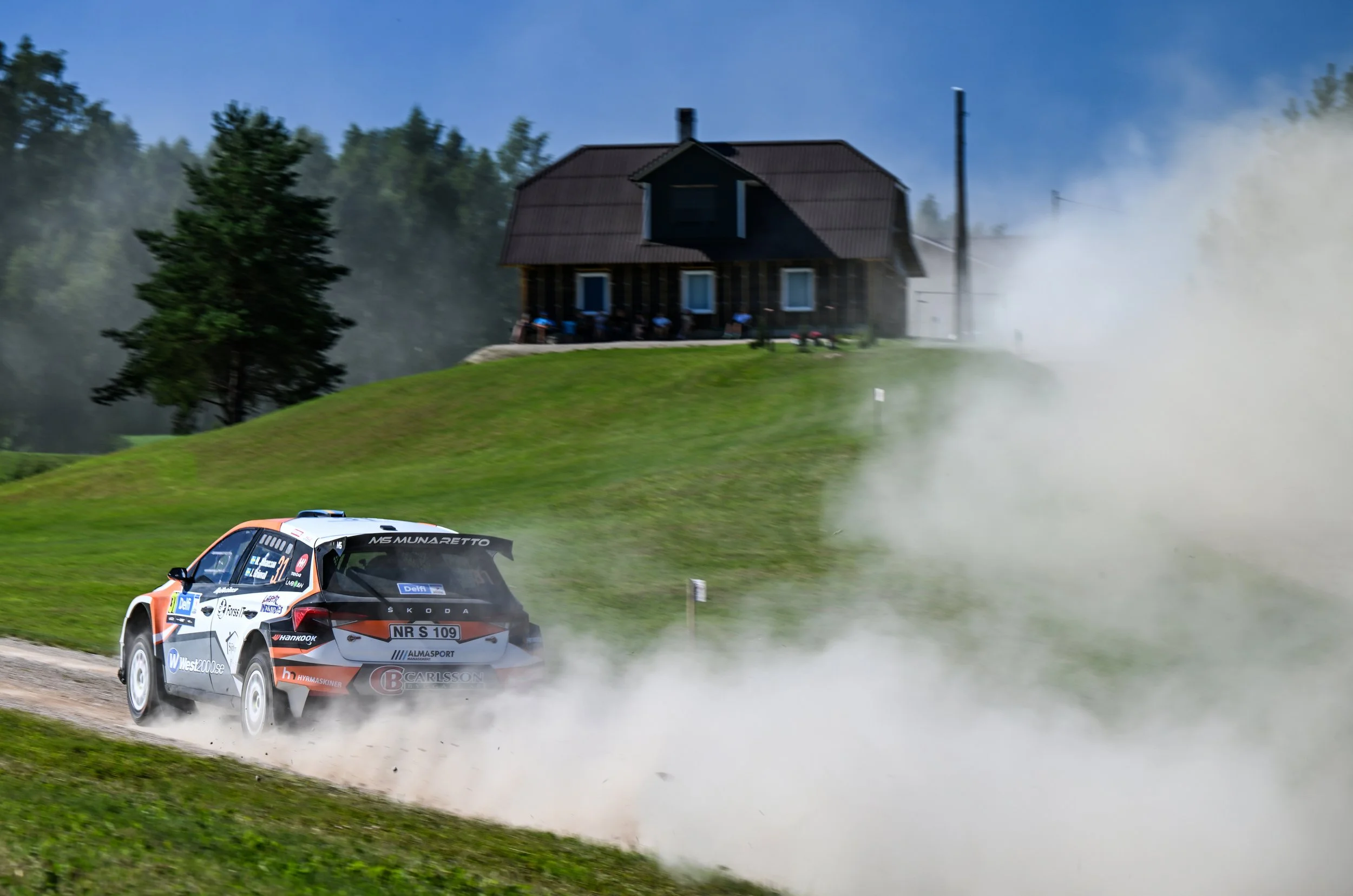A rally car speeding along a dirt road, kicking up dust and smoke, with a house and trees in the background.