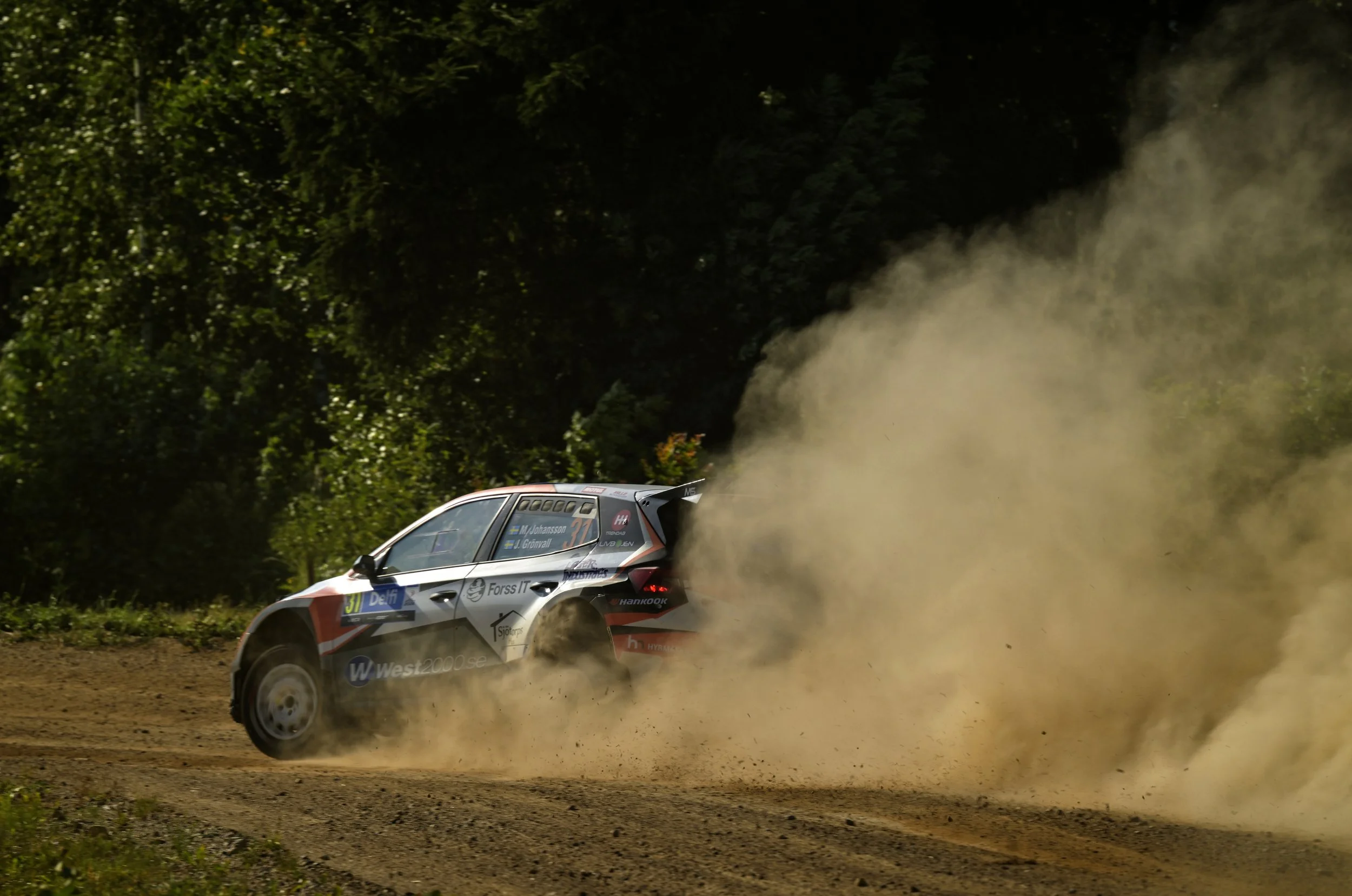 A rally car speeding on a dirt road kicking up dust, surrounded by green trees in the background.