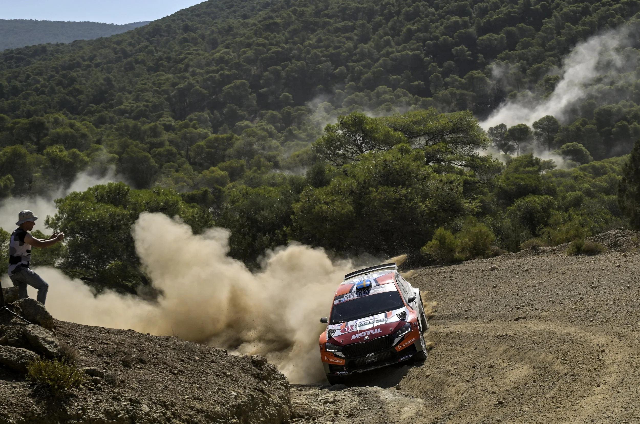 A rally car speeds along a dirt trail kicking up dust with a forested mountain in the background and a person taking a photo nearby.