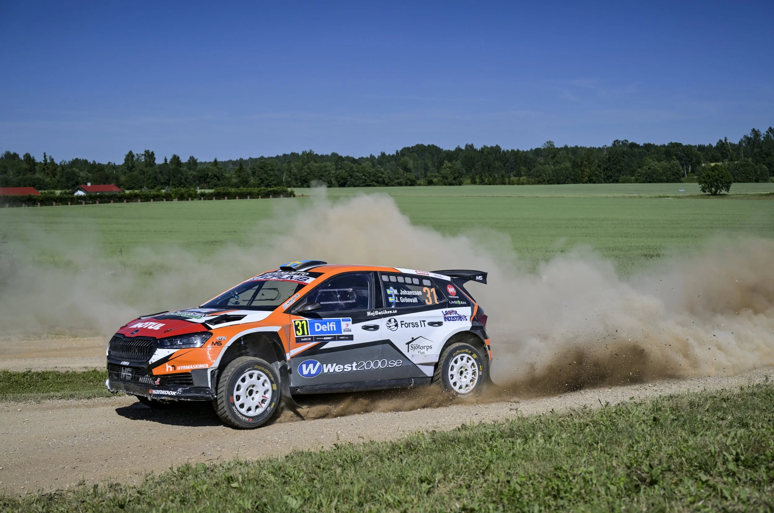 A rally car speeding on a dirt road, kicking up dust behind it, with a rural landscape of green fields, trees, a blue sky, and houses in the background.